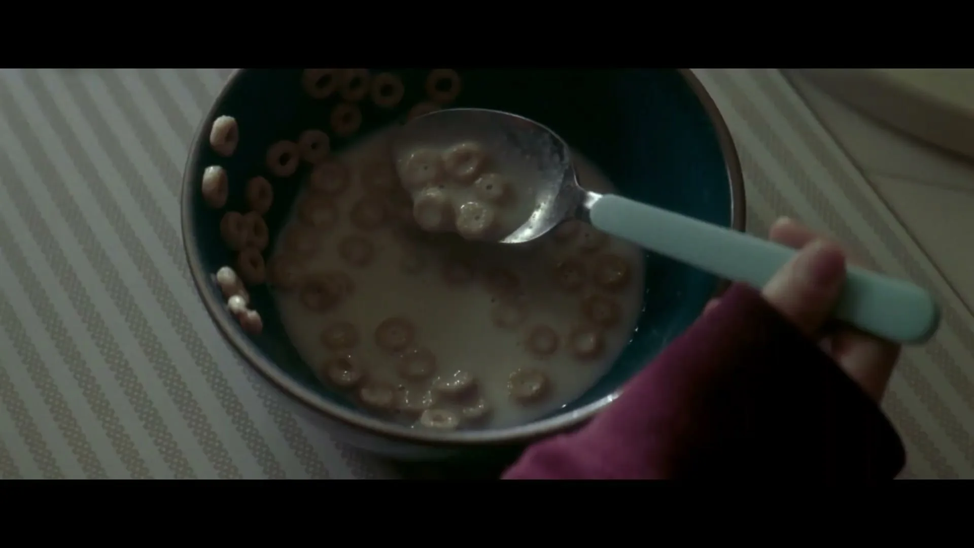 A child's hand in a purple sleeve holds a spoon in a bowl of cereal and milk on a striped placemat.