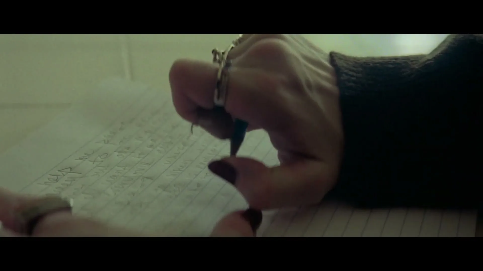 A person's hand with dark nail polish and rings writes "help me" on a lined piece of paper with a pen, in a dimly lit room.