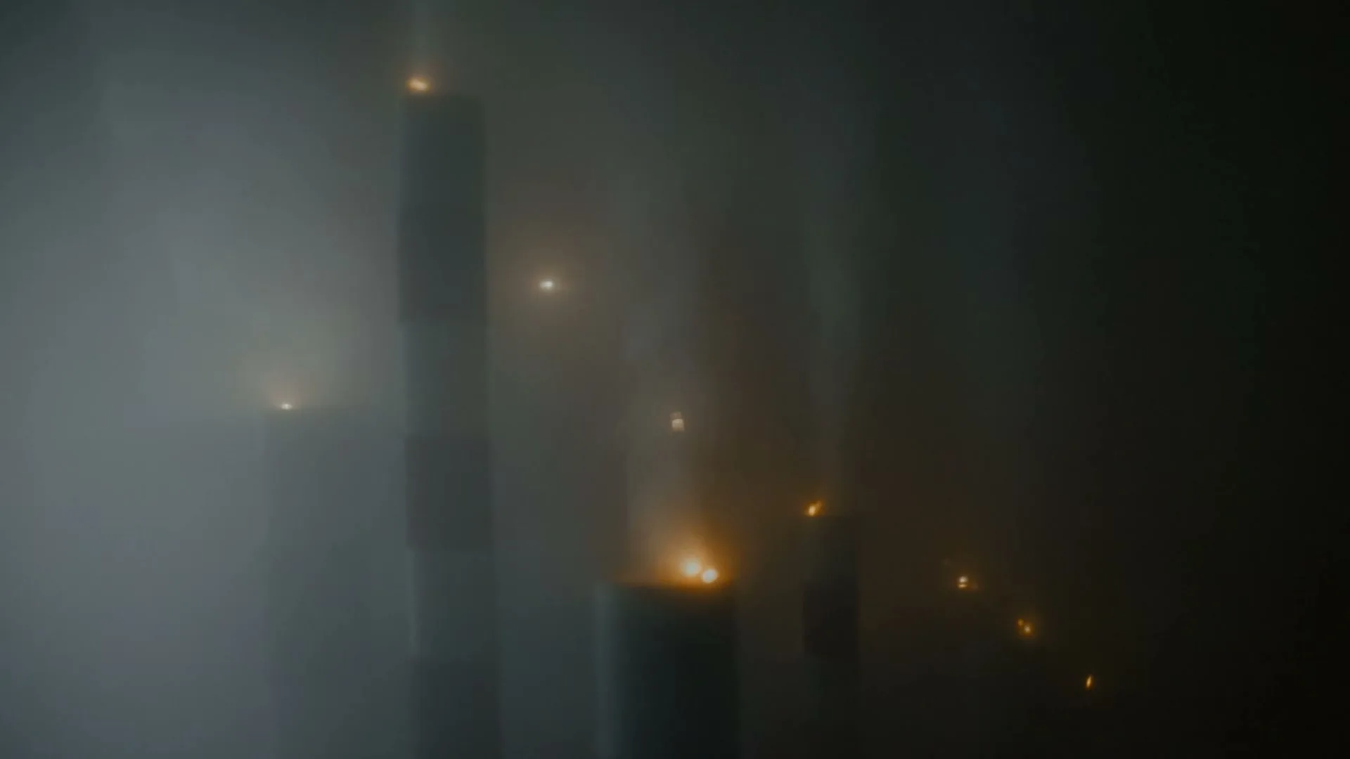 A wide shot of multiple industrial smokestacks emitting thick smoke, with small fires burning on top of some of them, creating a hazy, dark atmosphere.