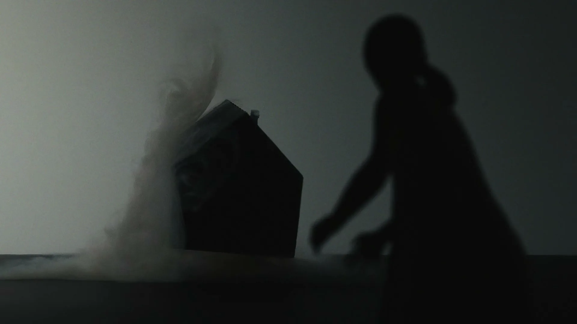 A child's silhouette is cast on a wall as a miniature house model is engulfed in smoke, then lifted by an unseen force.