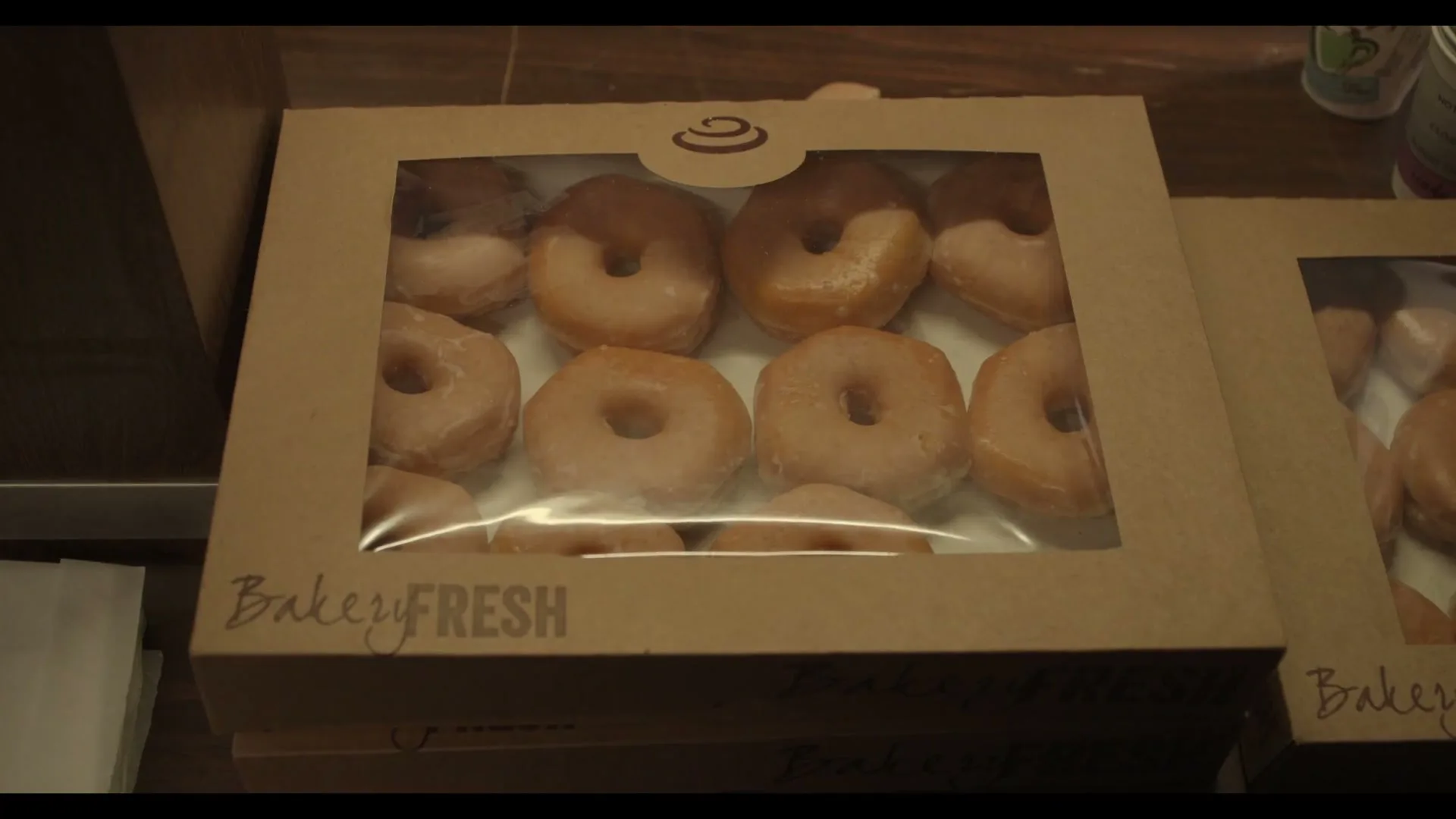 A box of glazed and chocolate-frosted donuts sits on a counter, with "BakeryFresh" printed on the side, in an interior setting.