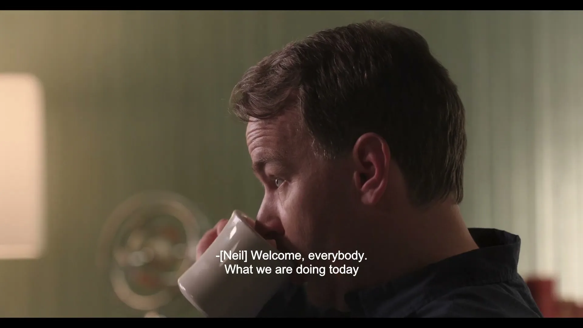 A man in his 30s, wearing a dark blue shirt, sips from a white mug in a softly lit room with a light green wall.