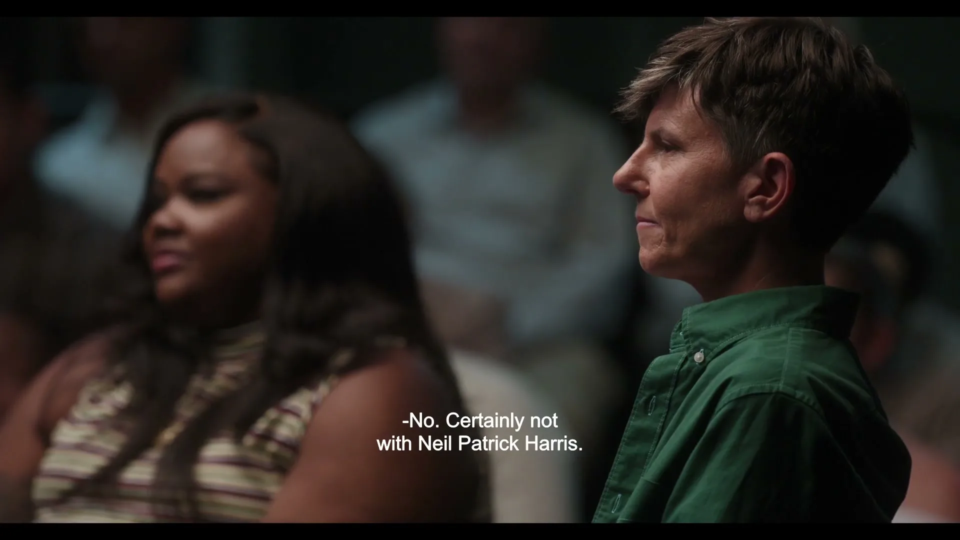 A person with short hair in a green shirt sits in profile, listening intently in a dimly lit group setting, with another person out of focus beside them.