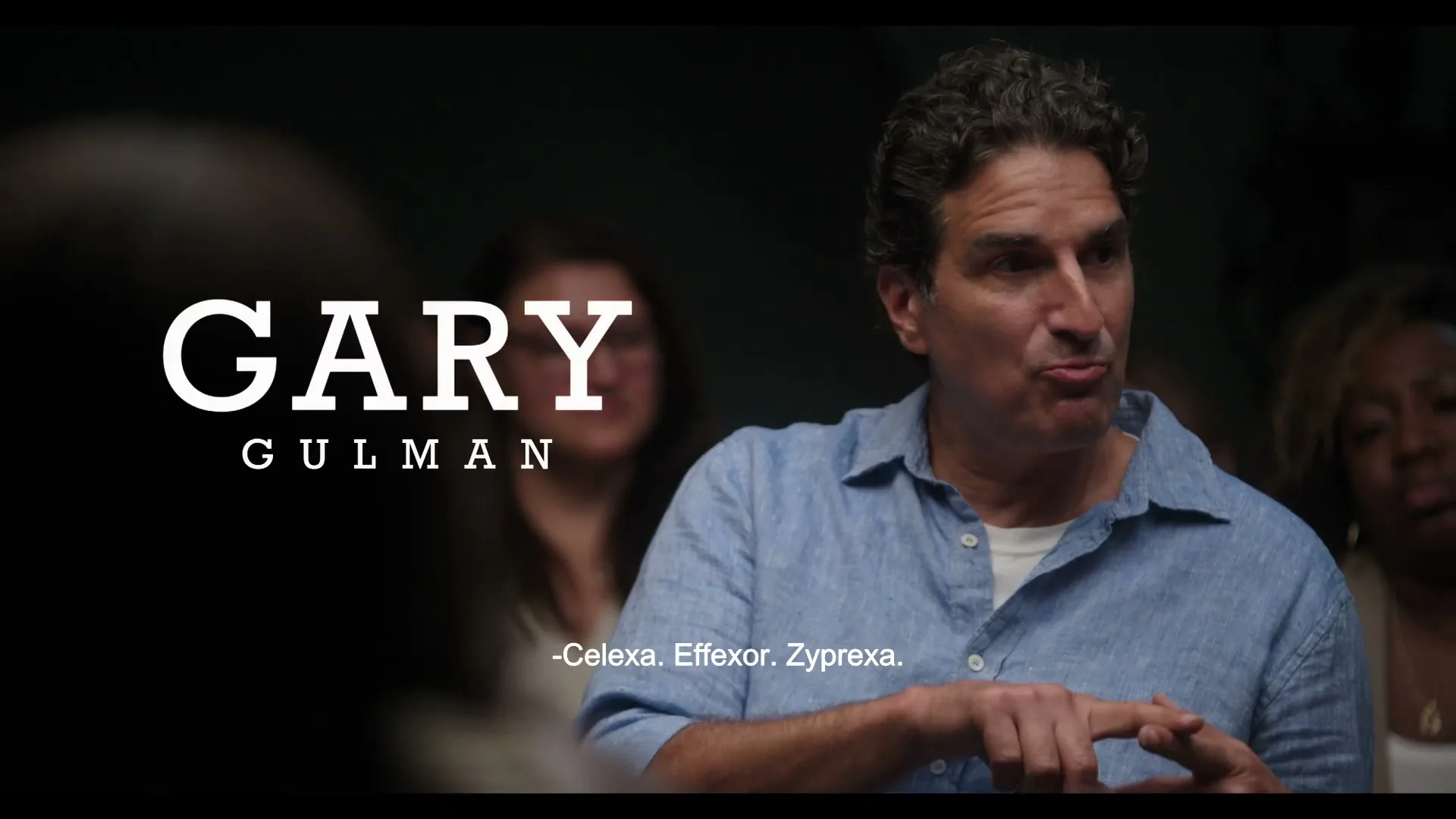 A male comedian in his 50s, wearing a blue shirt, speaks animatedly in a group therapy session, with his name displayed on screen.