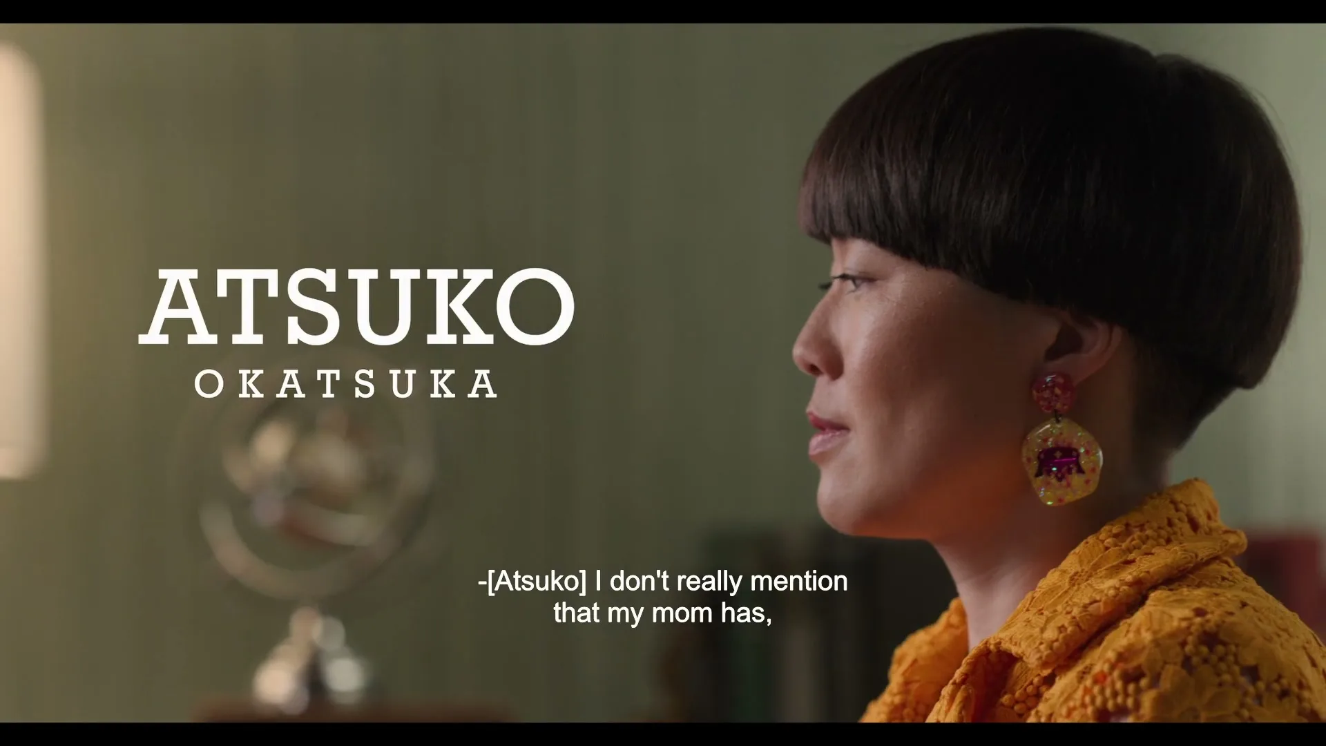 Atsuko Okatsuka, a woman with a bowl cut and yellow top, looks down while speaking in an interior setting.
