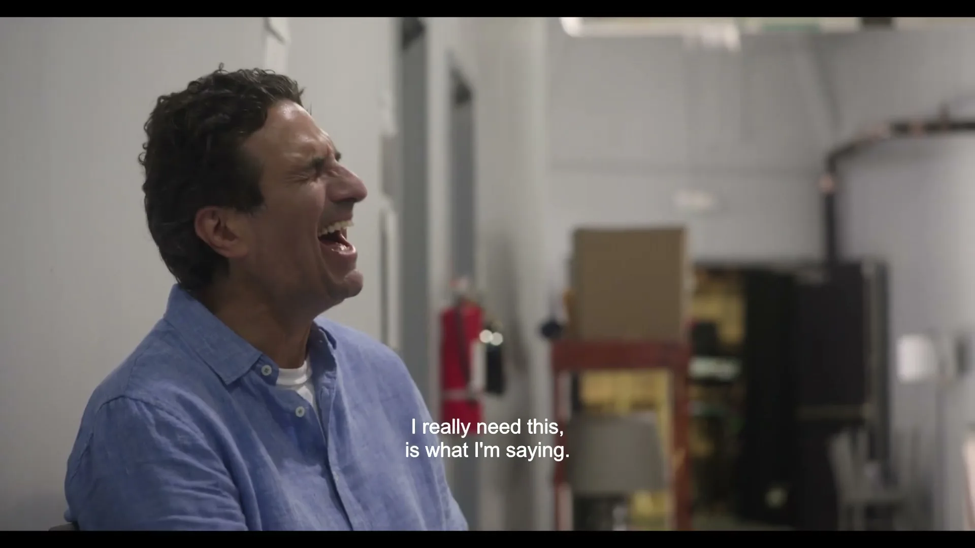 A man in his 40s, wearing a blue shirt, laughs heartily while sitting in an interior setting, with a fire extinguisher visible in the background.