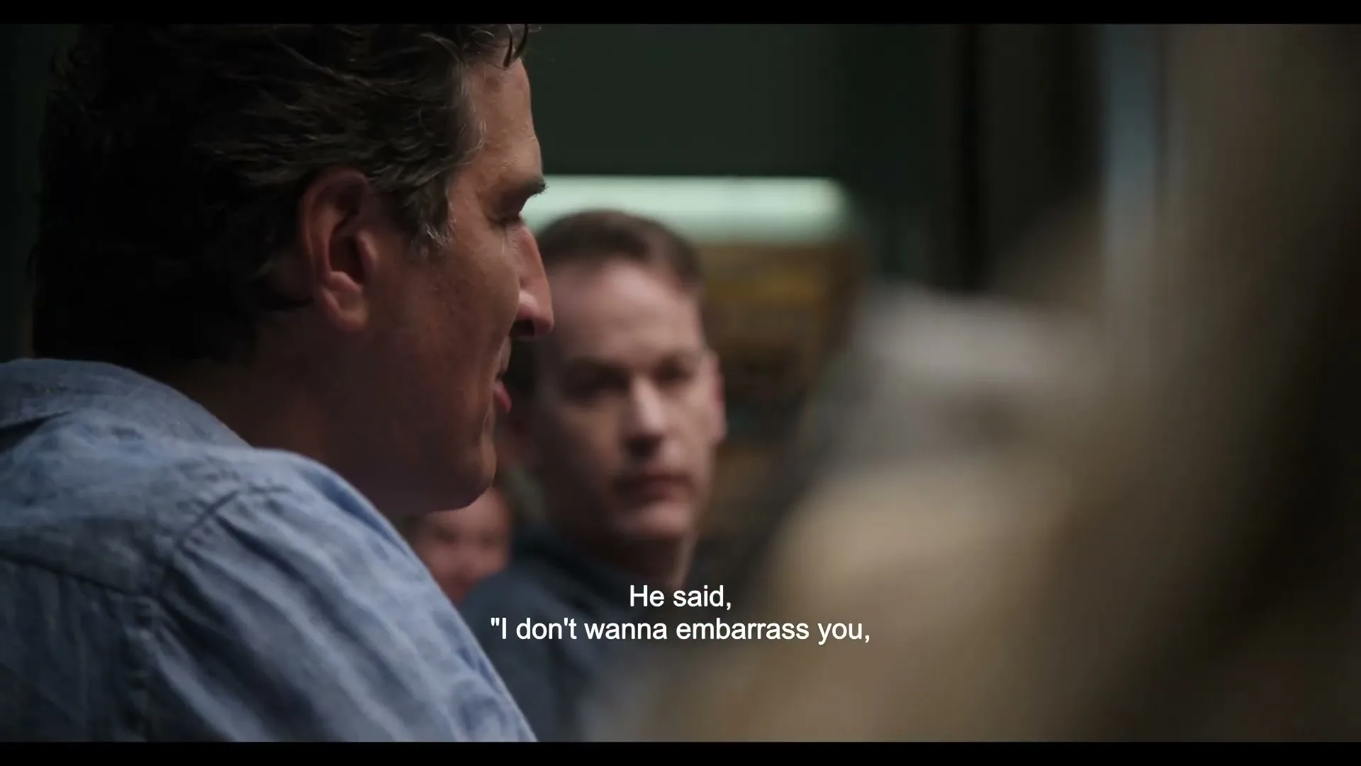 A man with dark, curly hair and a blue shirt speaks in profile, while another man listens intently in the background during a group therapy session.