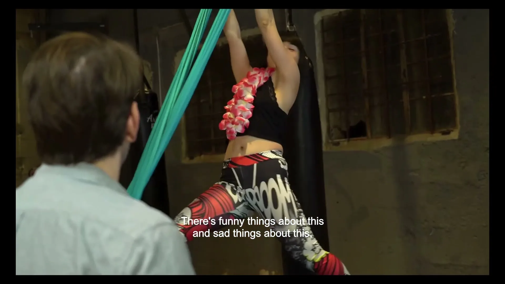 A woman in a black top and patterned leggings, wearing a lei, hangs from aerial silks, smiling at a man whose back is to the camera in a gym.
