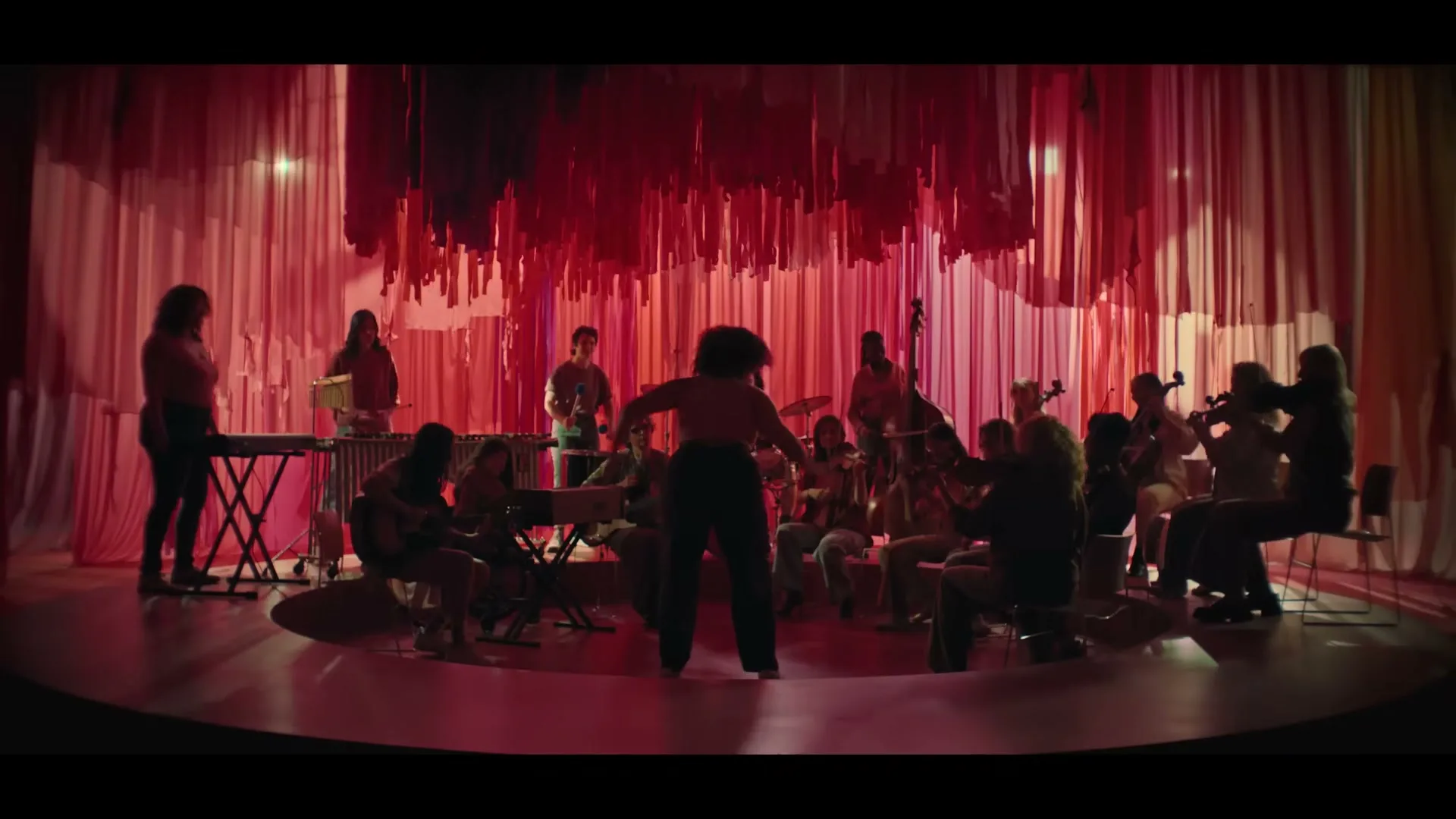 A diverse group of women and girls in a pink-red room with a conductor in the center, playing various instruments.
