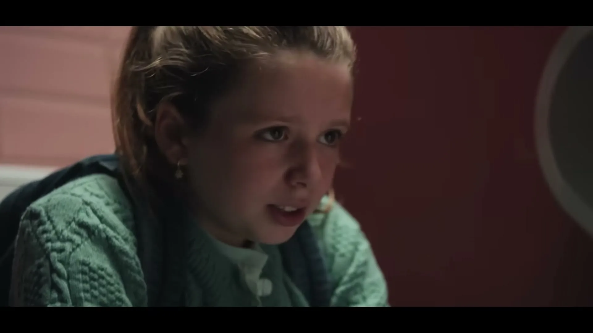 A young girl with light brown hair in a green sweater looks up with a worried expression in a pink-tiled bathroom.
