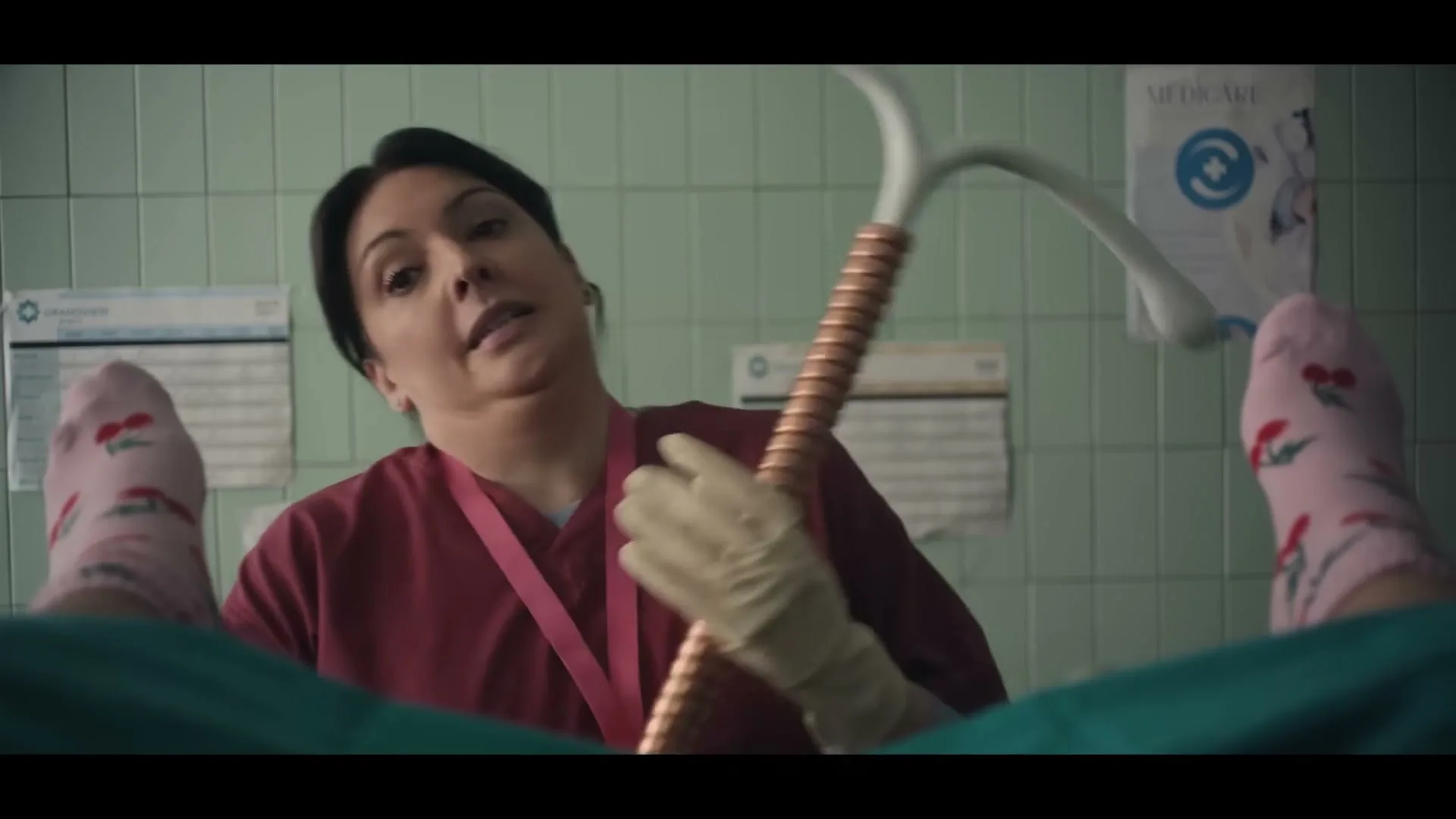 A female doctor in red scrubs and gloves holds up a speculum-like object, looking at the camera, with a patient's feet in pink cherry socks visible in the foreground.