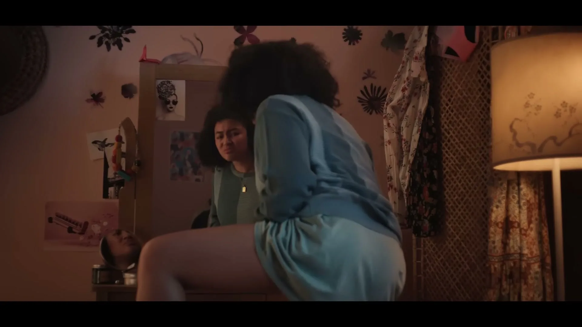 A young woman with curly hair sits on a stool, looking at her reflection in a mirror in a dimly lit bedroom.