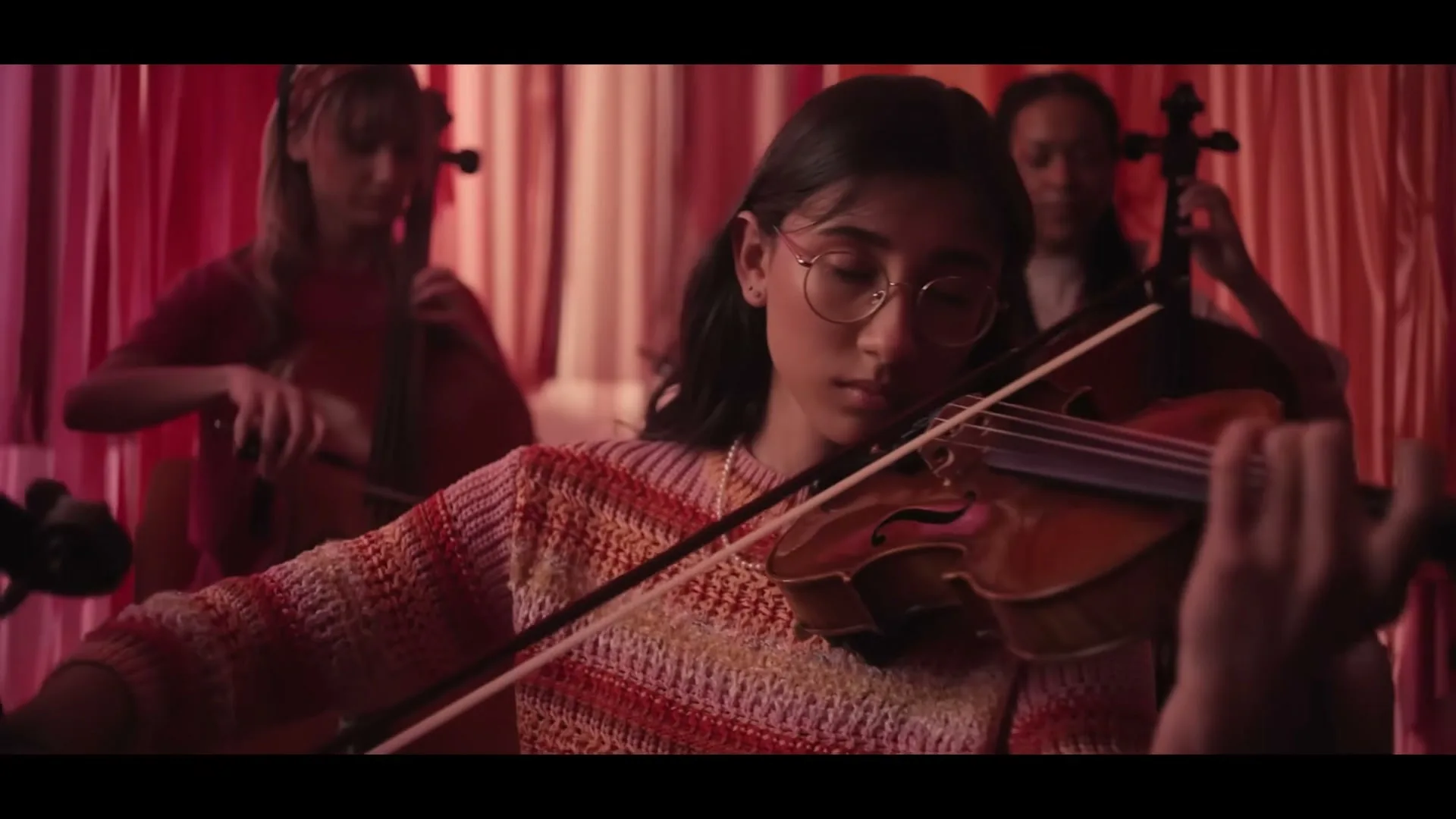 A young woman with glasses plays the violin with intense focus, surrounded by other musicians and red draped fabric in a warm, intimate setting.