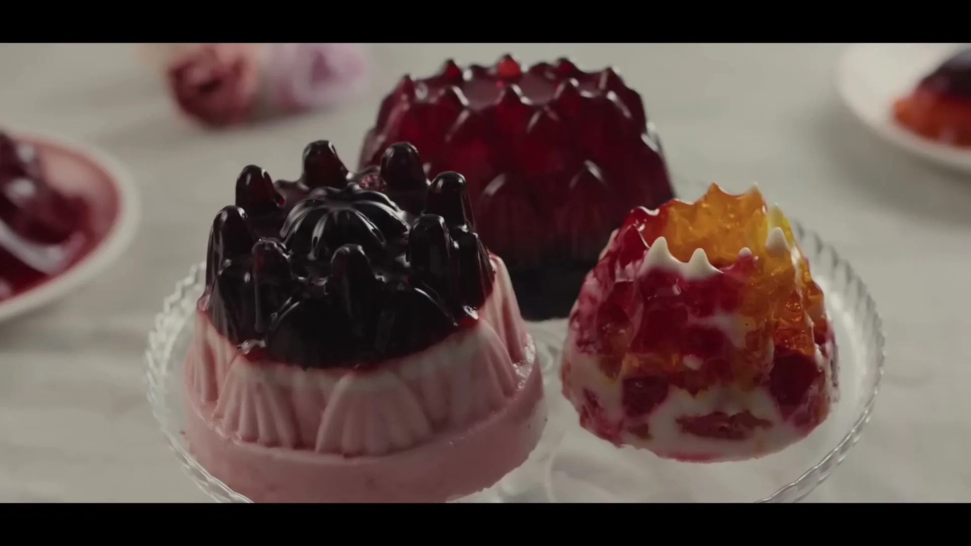 Three colorful, molded gelatin desserts, one pink with dark topping, one red, and one orange and white, are displayed on a glass plate.