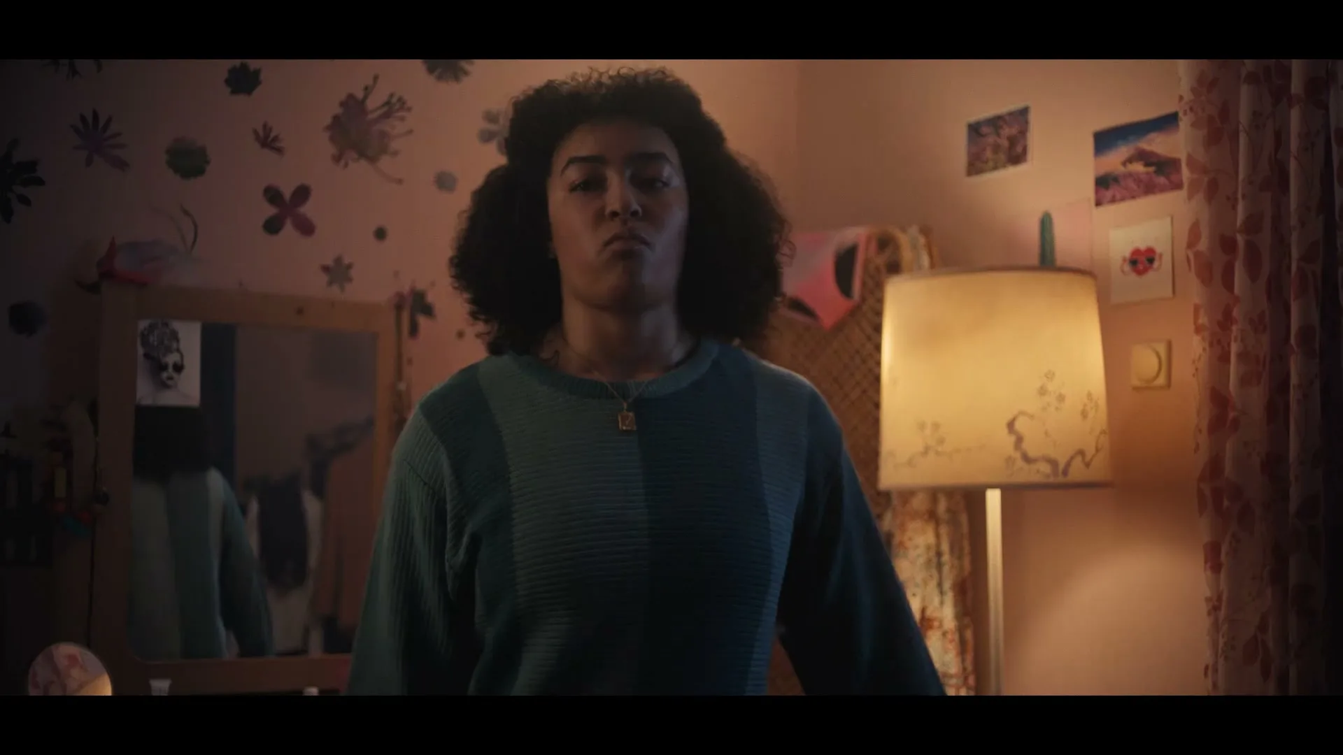 A young woman with curly hair, wearing a blue sweater, stands in her bedroom, looking directly at the camera with a serious expression.