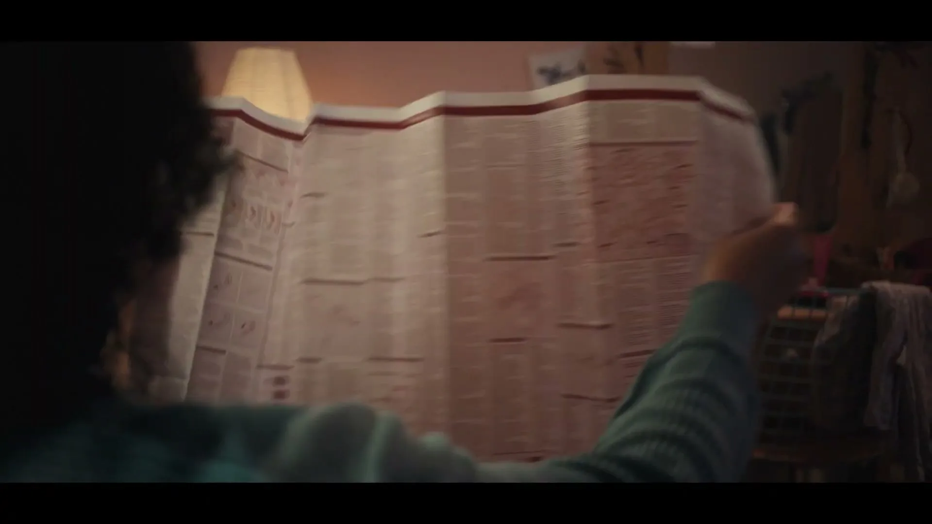 A young person with curly hair unfolds a large, detailed instruction manual in a dimly lit bedroom.