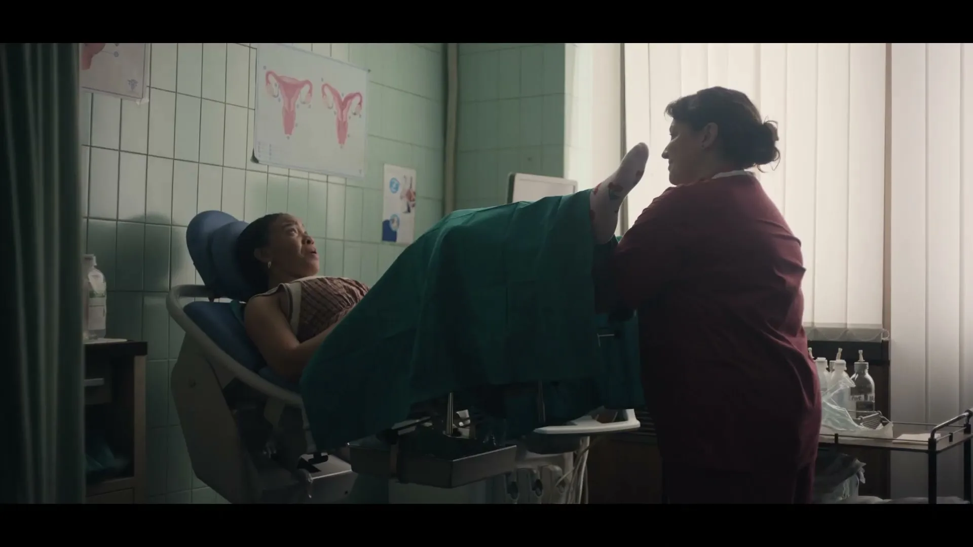 A woman in a medical gown lies on an examination table while a female doctor in scrubs performs a gynecological exam in a clinic.