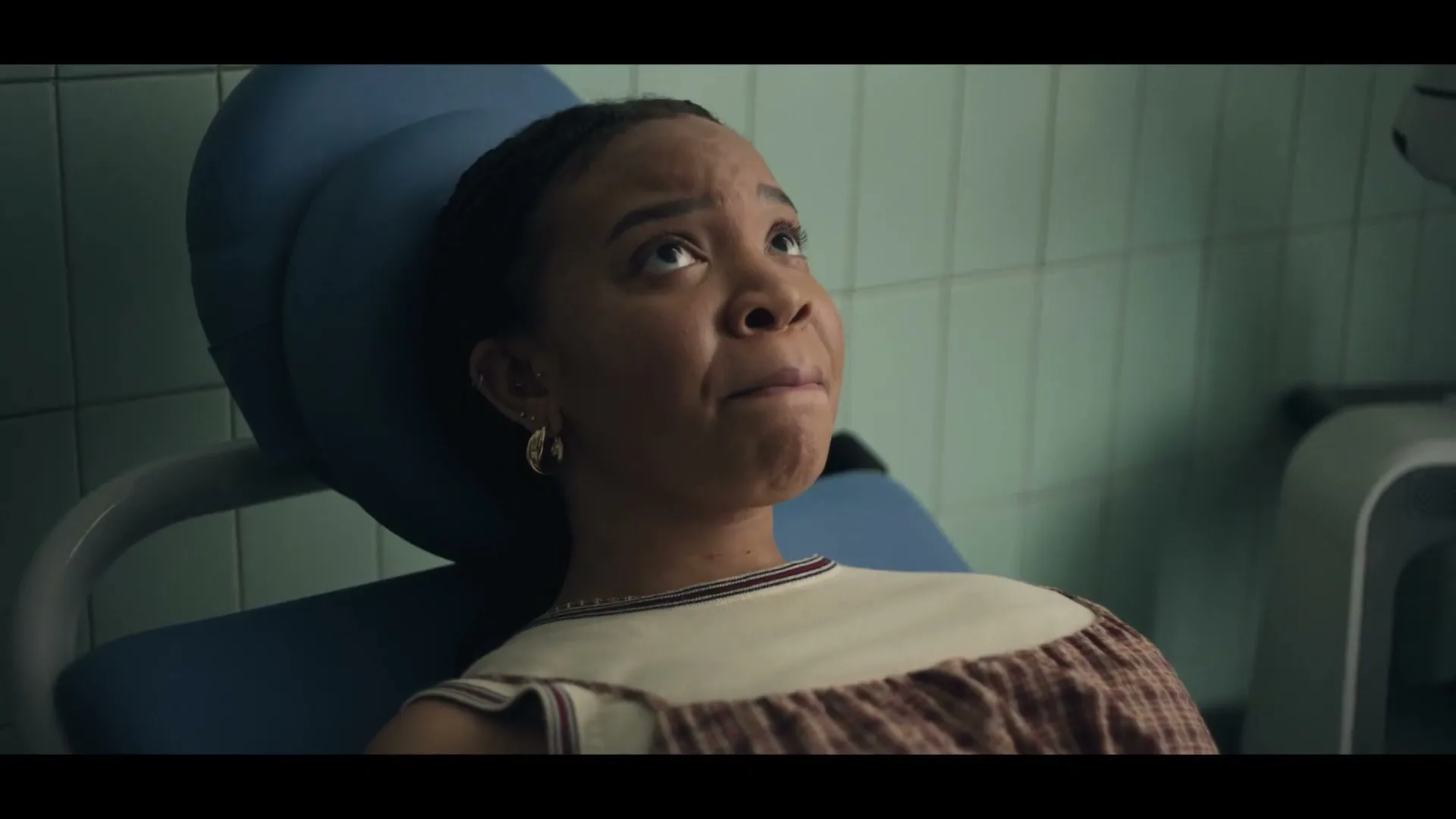 A young woman with dark hair and a patterned dress sits in a blue dental chair, looking up with a slightly concerned expression in a dentist's office.