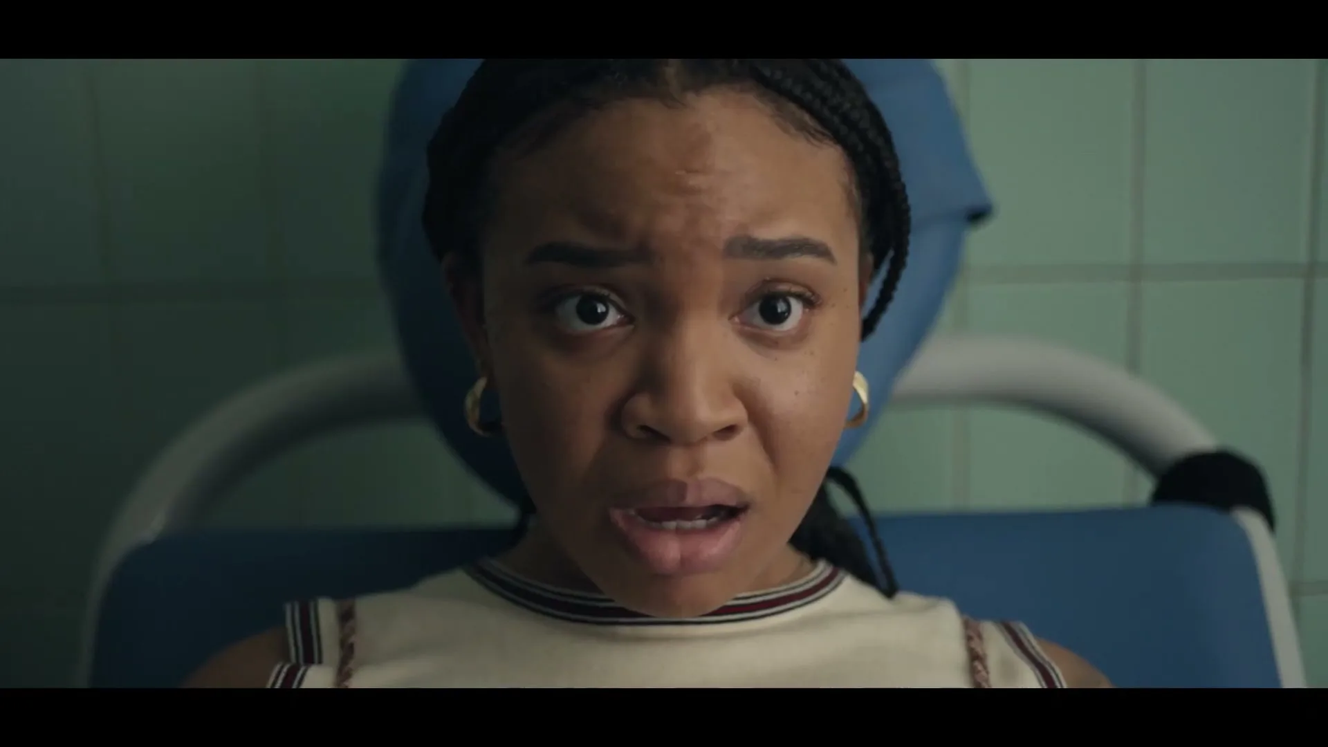 A young Black woman with braided hair and gold hoop earrings looks directly at the camera with a surprised expression, sitting in a blue medical chair.