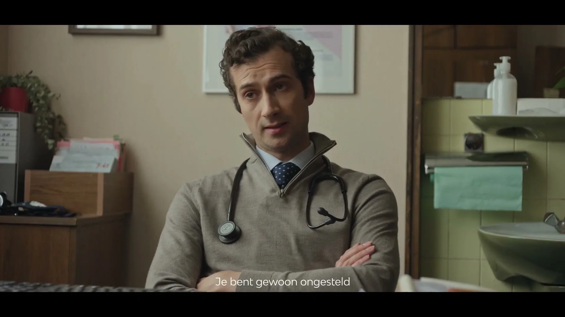A male doctor with a stethoscope around his neck sits at a desk, looking directly at the camera with a neutral expression in his office.