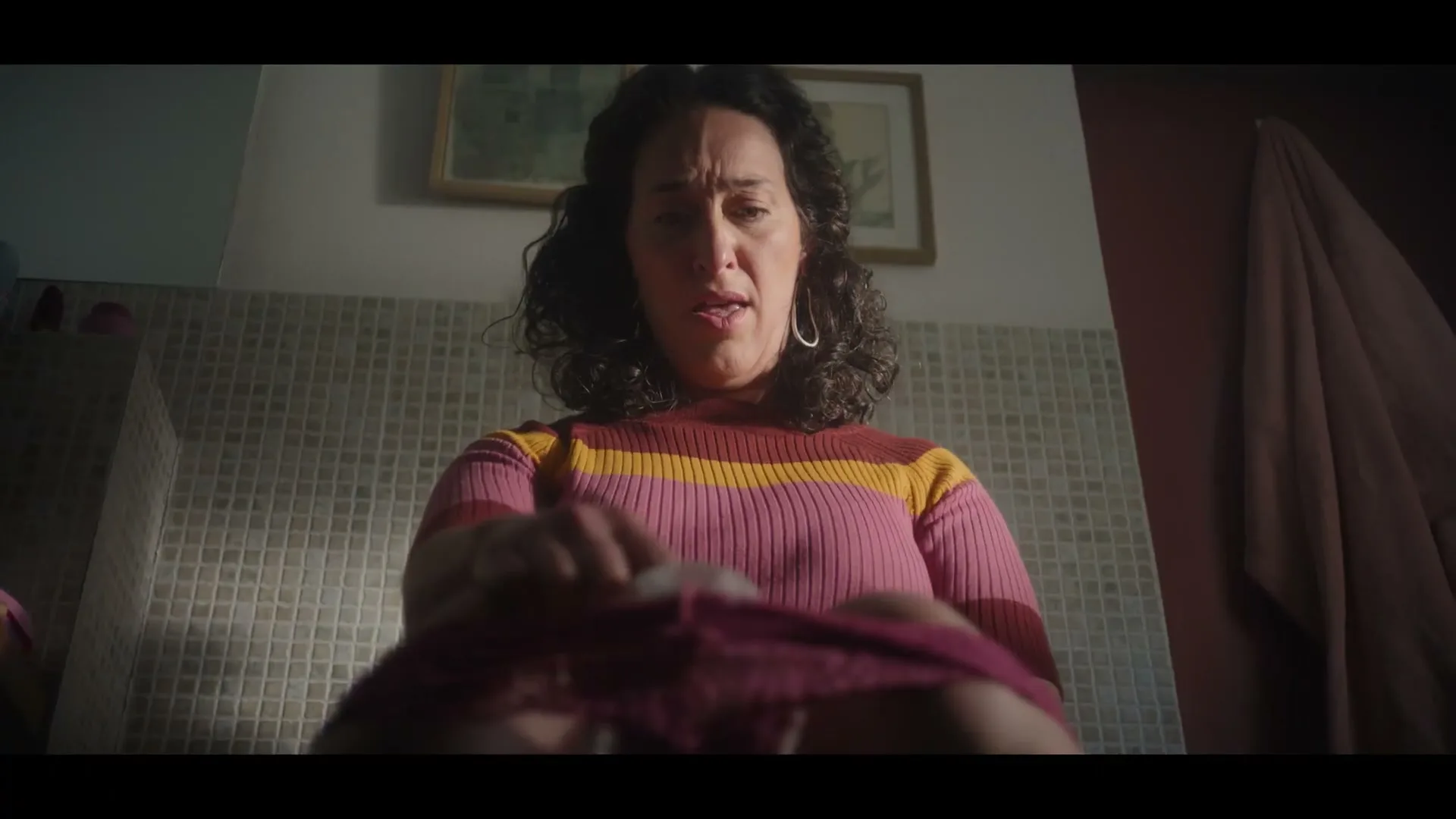 A woman with curly hair and a striped sweater sits on a toilet, looking down at her underwear with a concerned expression in a bathroom.