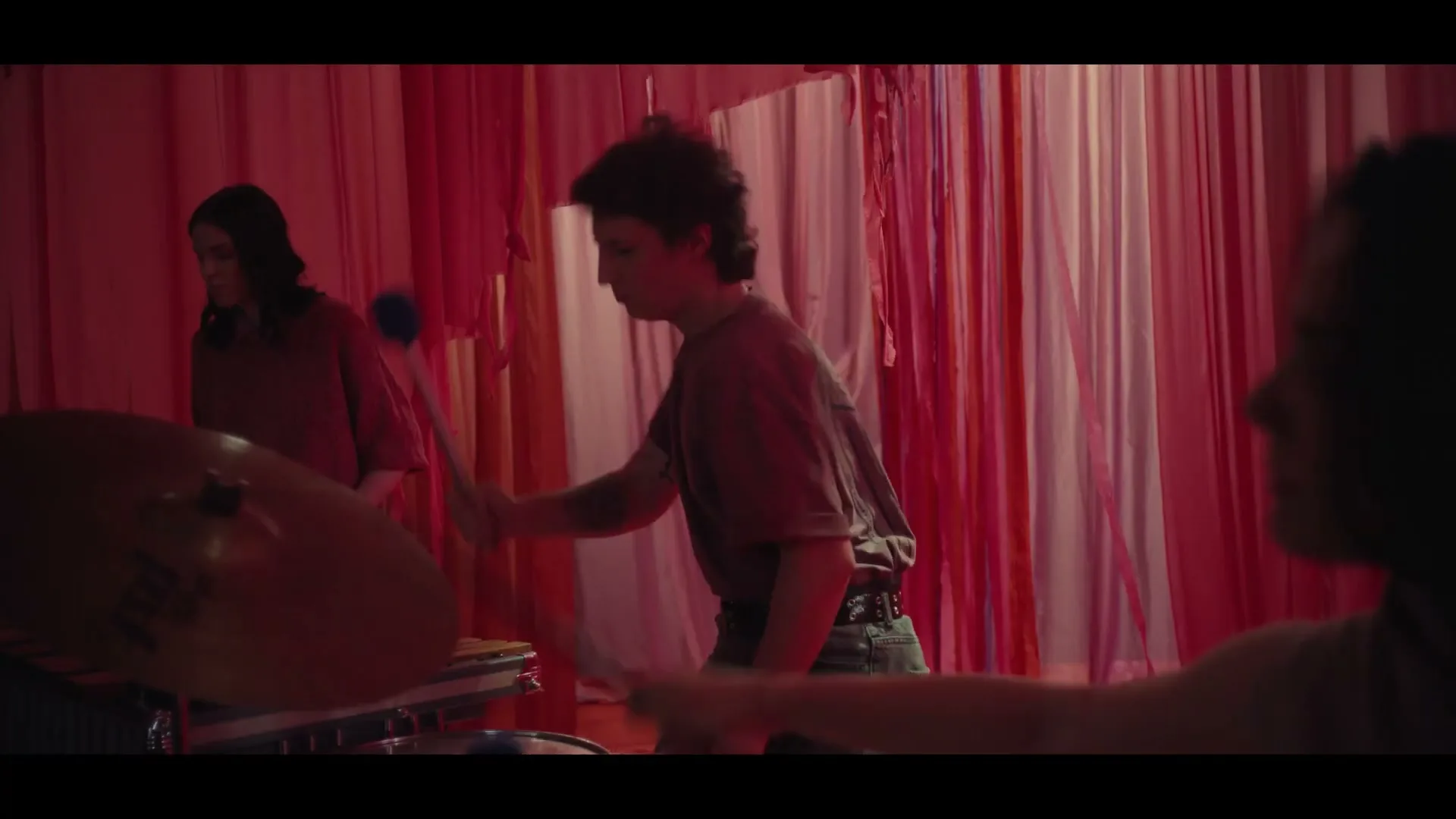 A young person with curly hair plays a vibraphone with mallets, while another person stands behind them, in a room with red fabric strips.