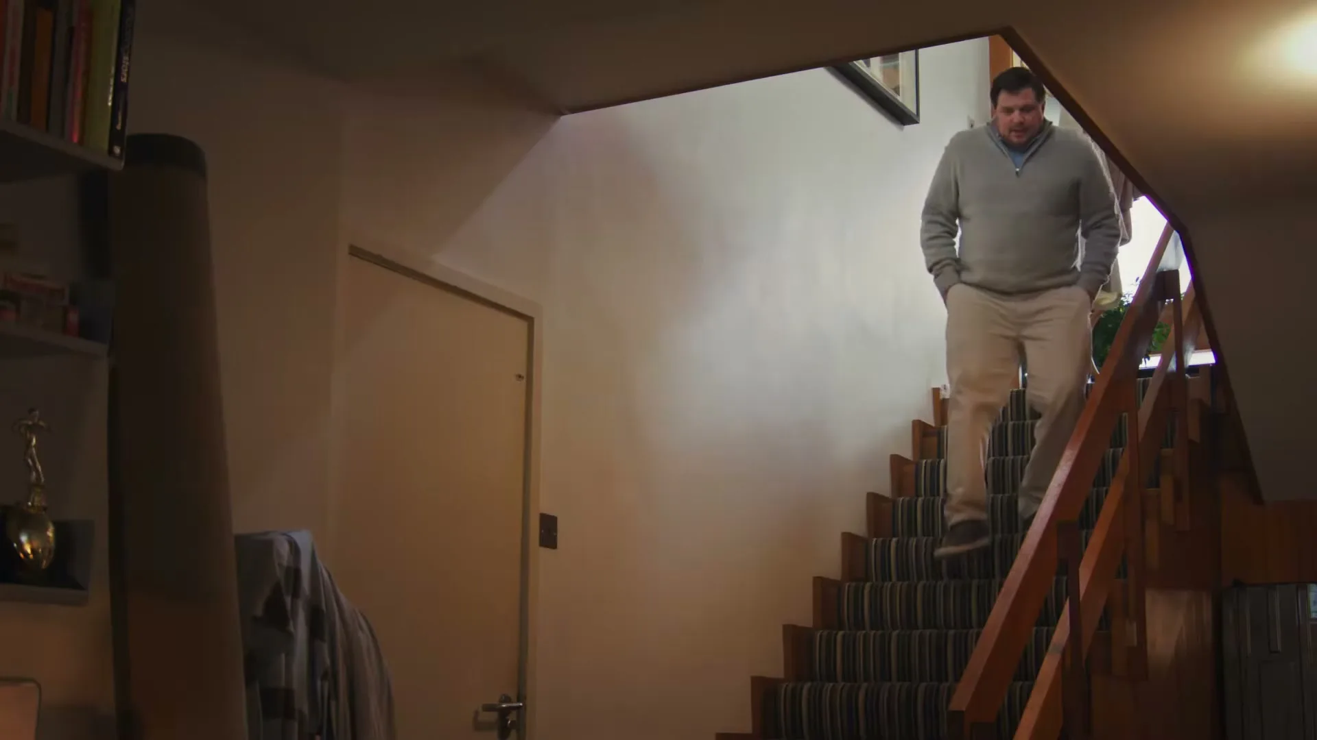 A man in his 40s walks down a carpeted staircase in a house, looking around with a concerned expression.