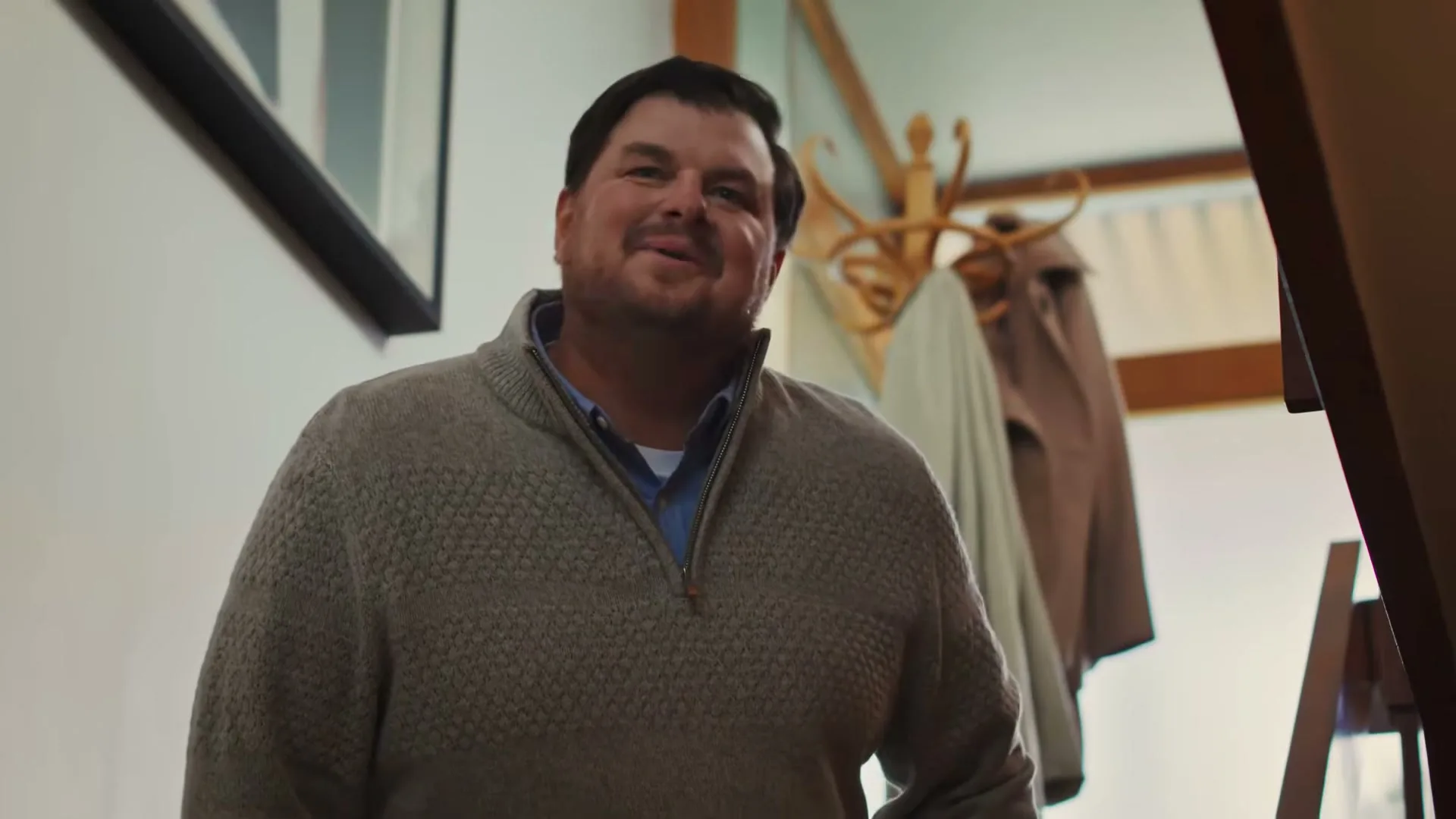 A man in a grey sweater smiles and looks up, standing in a bright entryway with a coat rack behind him.