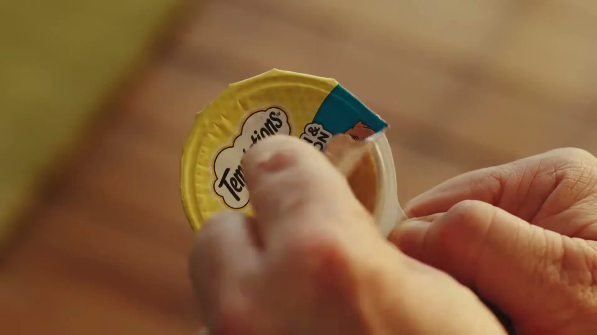 A person's hands peel back the lid of a Temptations cat food container, revealing two different colored cat food purees inside.