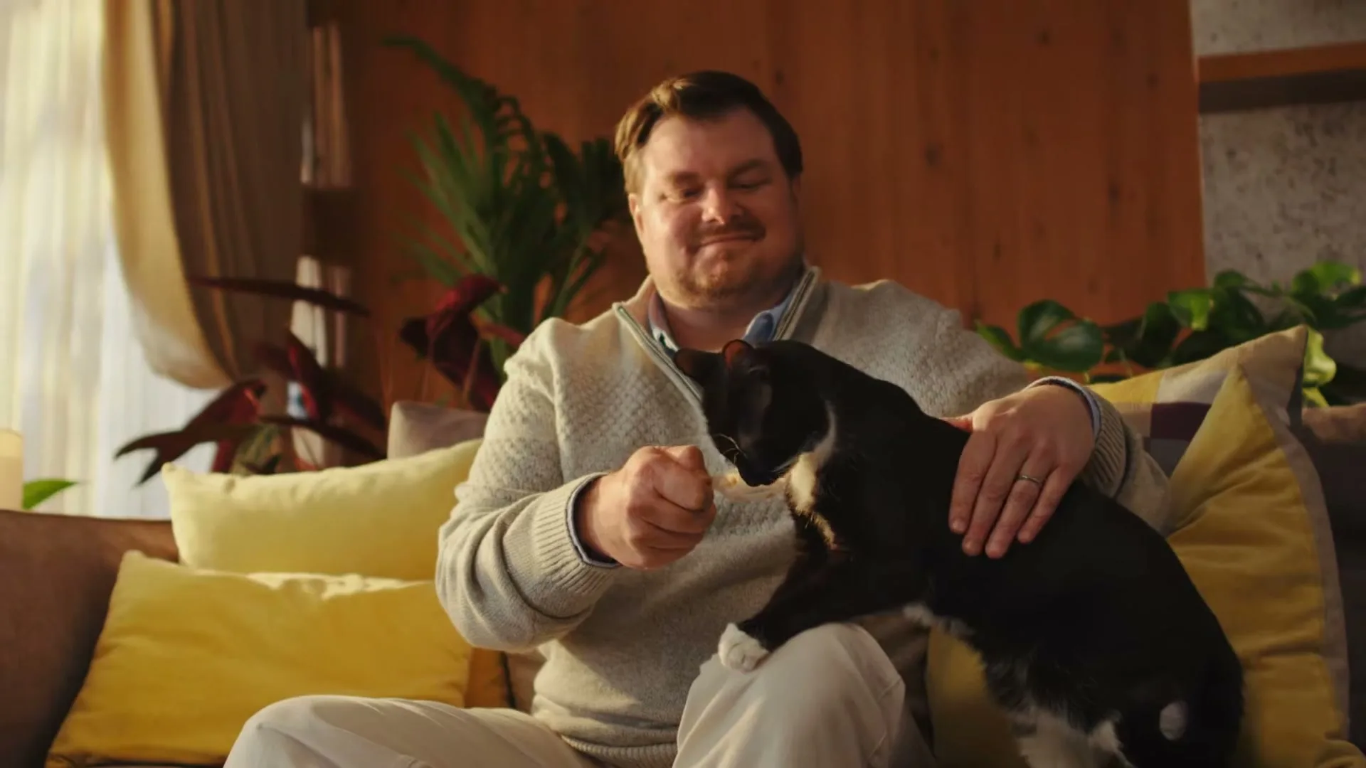 A man in a sweater sits on a couch, smiling as he feeds a black and white cat a treat from a small spoon.