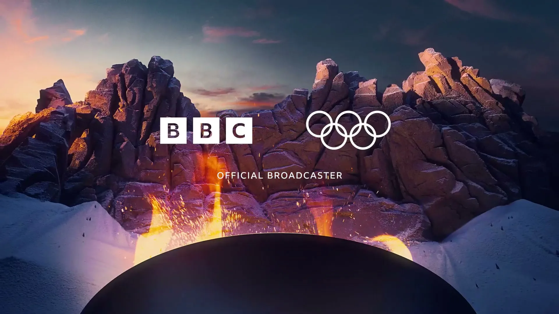 A wide shot of a snowy mountain landscape with a large fire pit in the foreground, featuring the BBC and Olympic rings logos, and the text "OFFICIAL BROADCASTER."