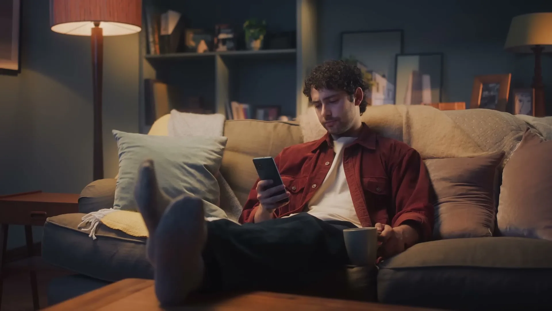 A young man with curly hair sits on a sofa, looking at his phone and holding a mug in a dimly lit living room.