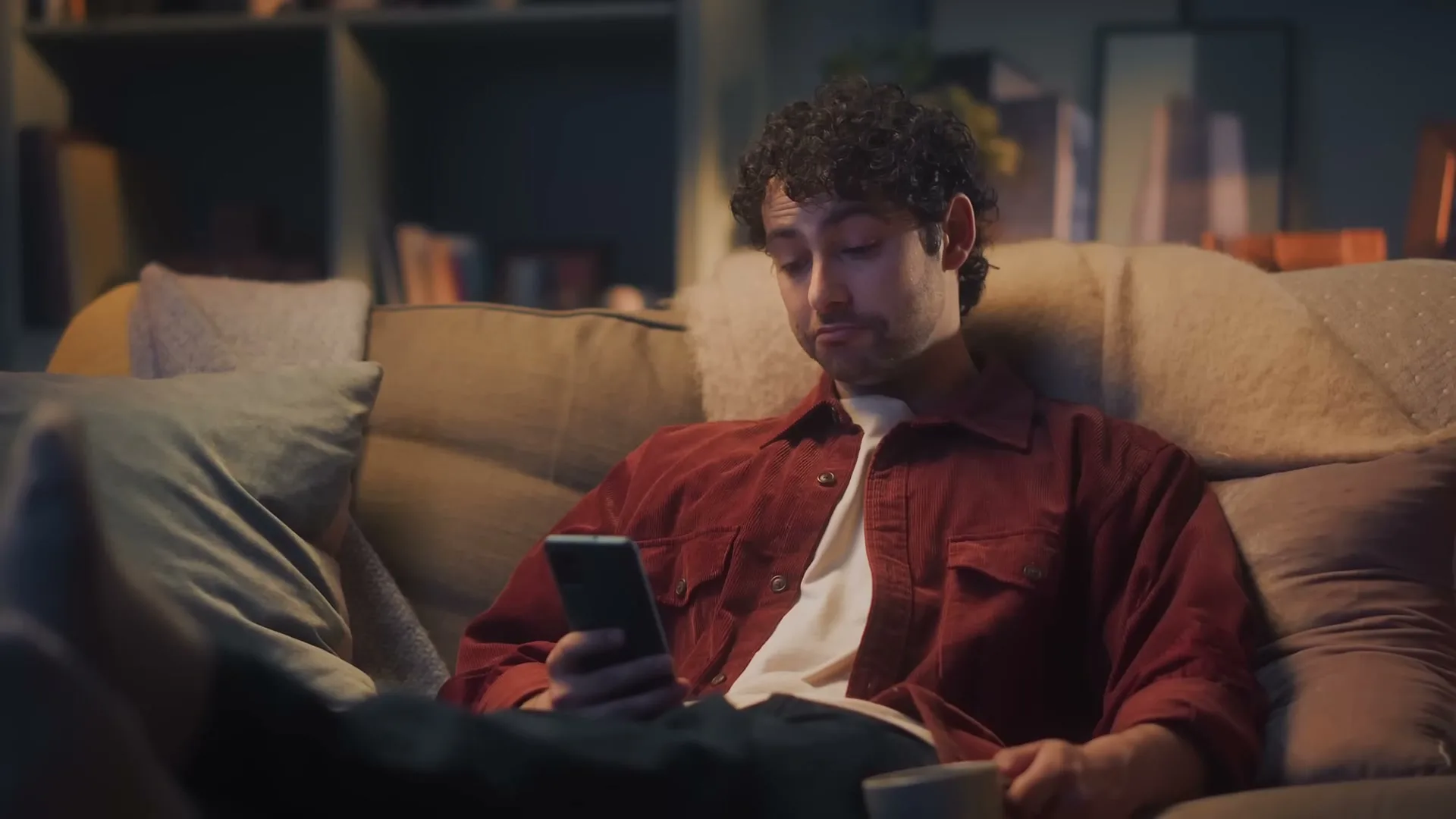 A young man with curly hair, wearing a red shirt, sits on a sofa in a dimly lit living room, looking at his phone.