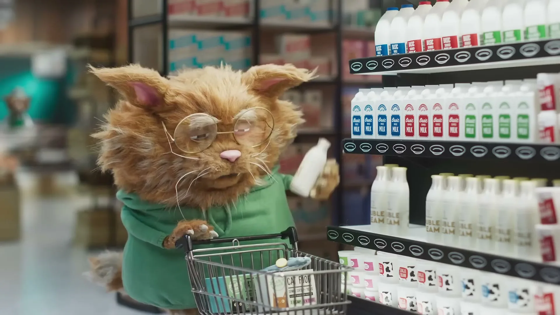 A fluffy orange cat wearing glasses and a green hoodie pushes a shopping cart down a supermarket aisle, looking at a small bottle of milk.