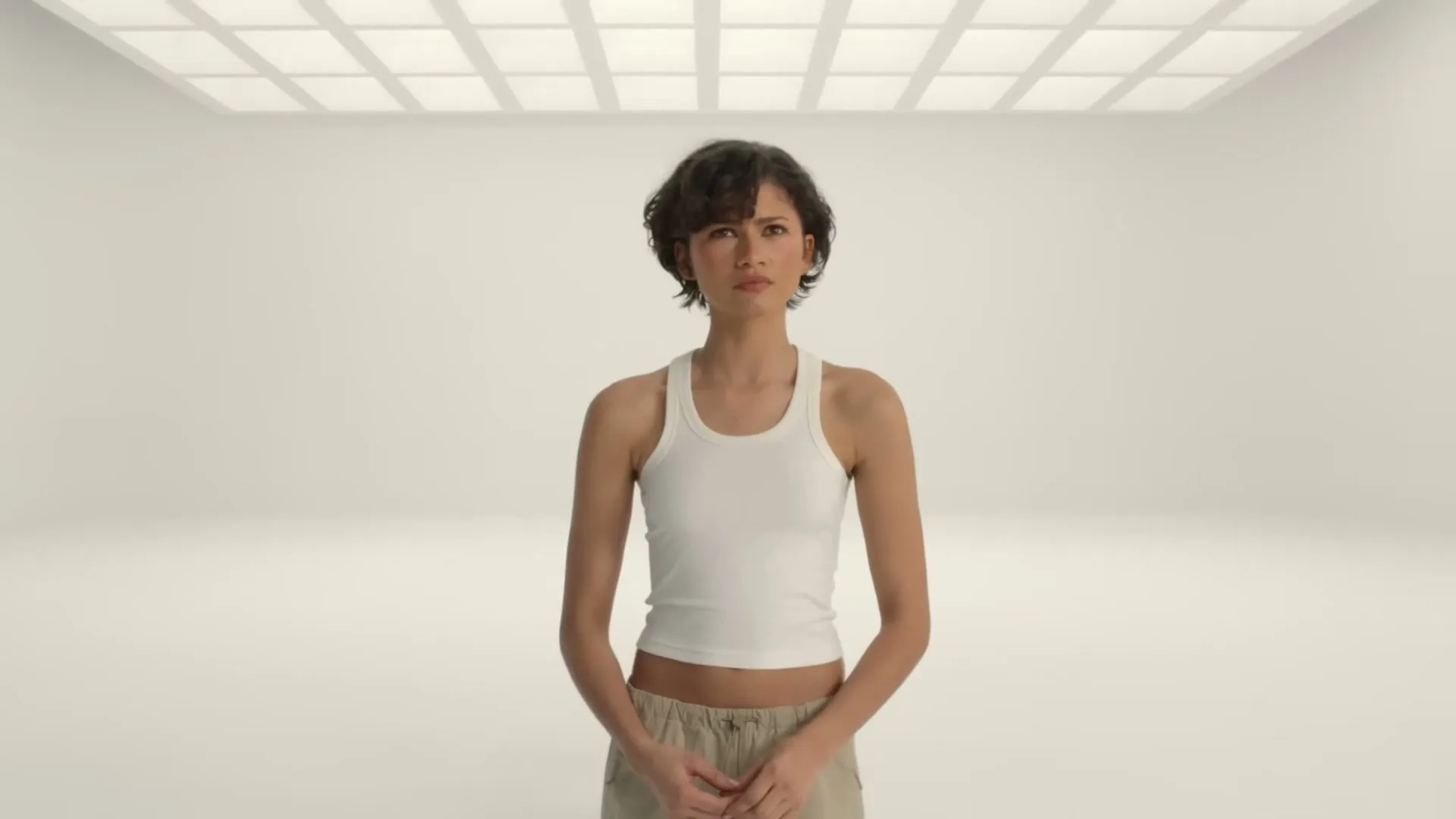 A young woman with short curly hair and a white tank top stands in a white studio, looking around as other dancers appear and disappear around her.