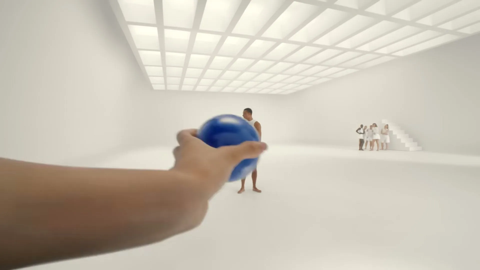 A hand throws a blue ball at a man in white shorts, who then inflates into a giant blue ball in a white room.