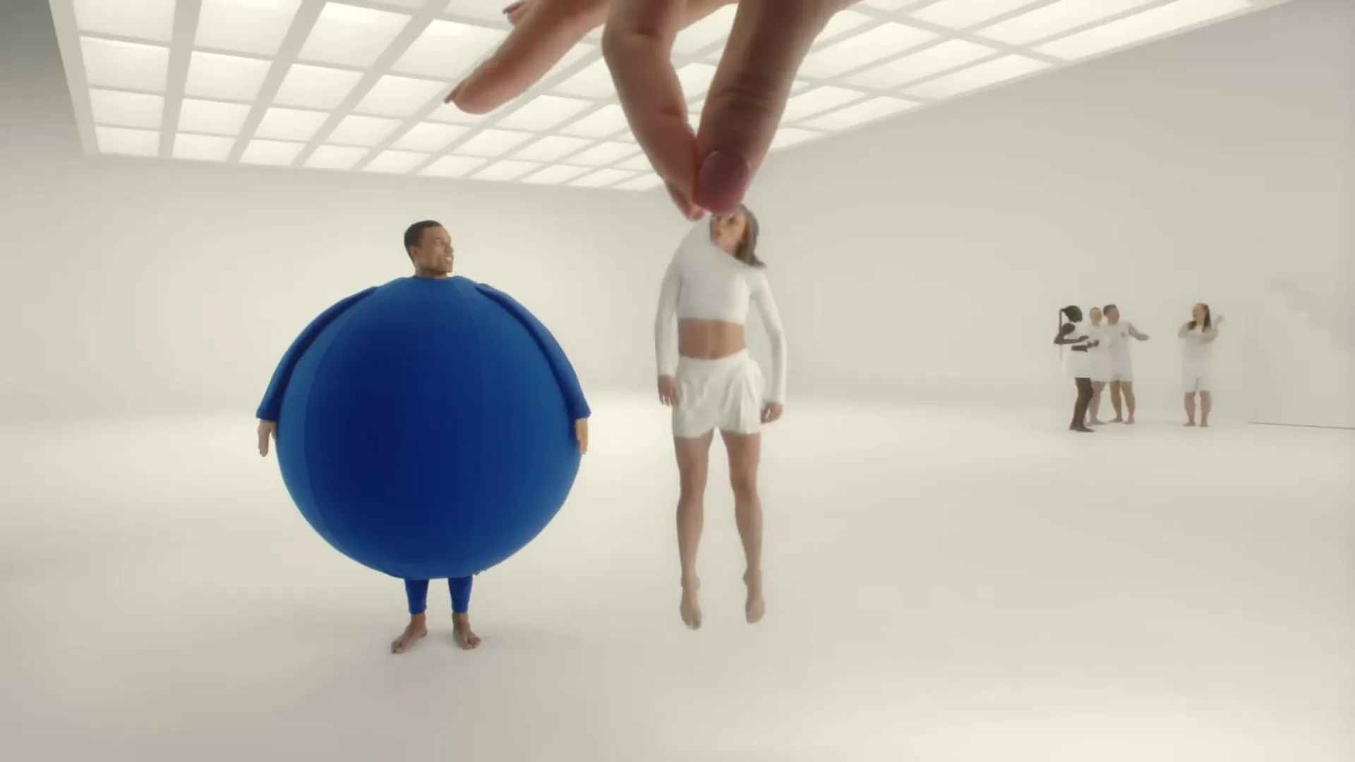 A man in a blue inflatable suit stands in a white studio as a giant hand drops a woman, who then inflates into a blue and white suit.