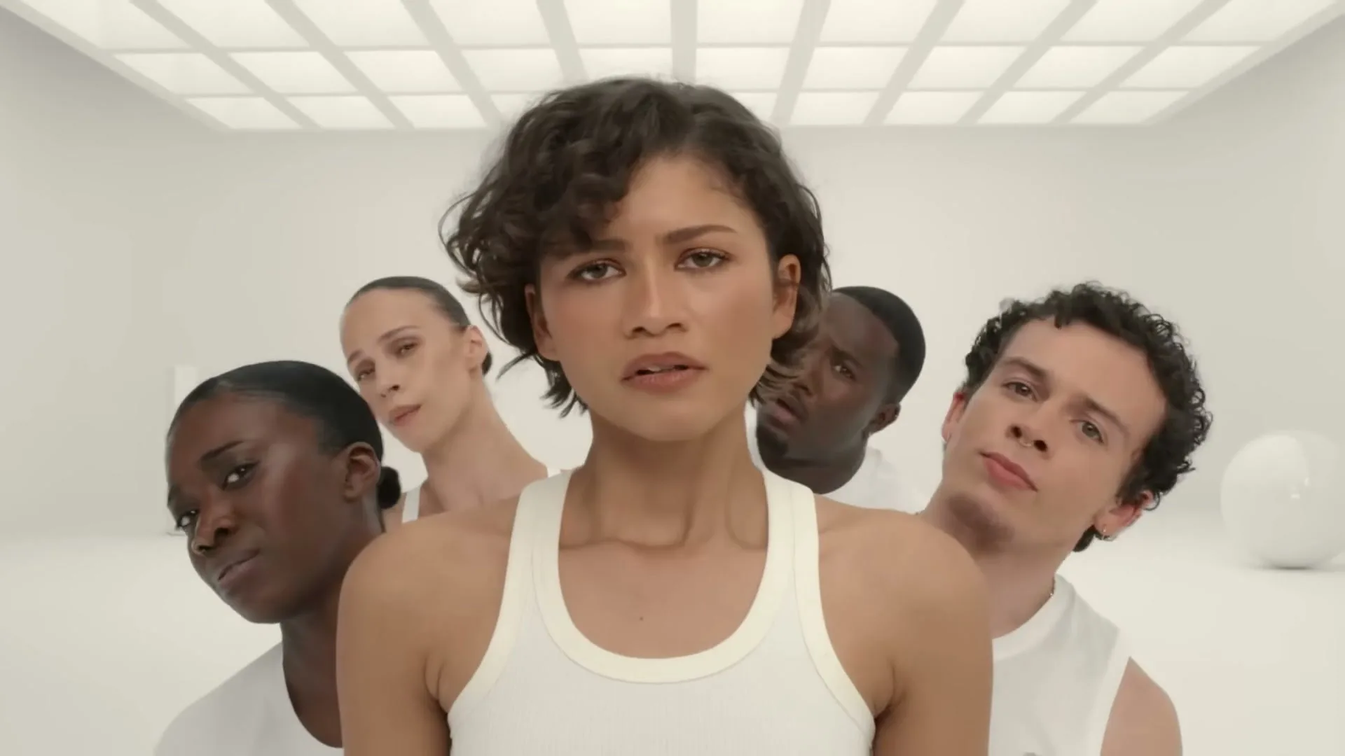 A young woman with short curly hair smiles, then looks confused and annoyed as four other people appear behind her in a white room.