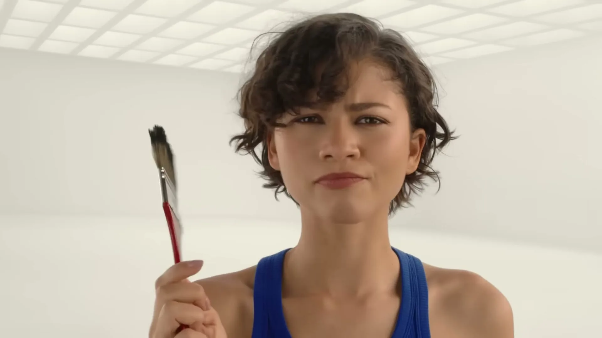 A young woman with short curly hair, wearing a blue tank top, holds a paintbrush and looks at it with a curious expression in a bright, white room.