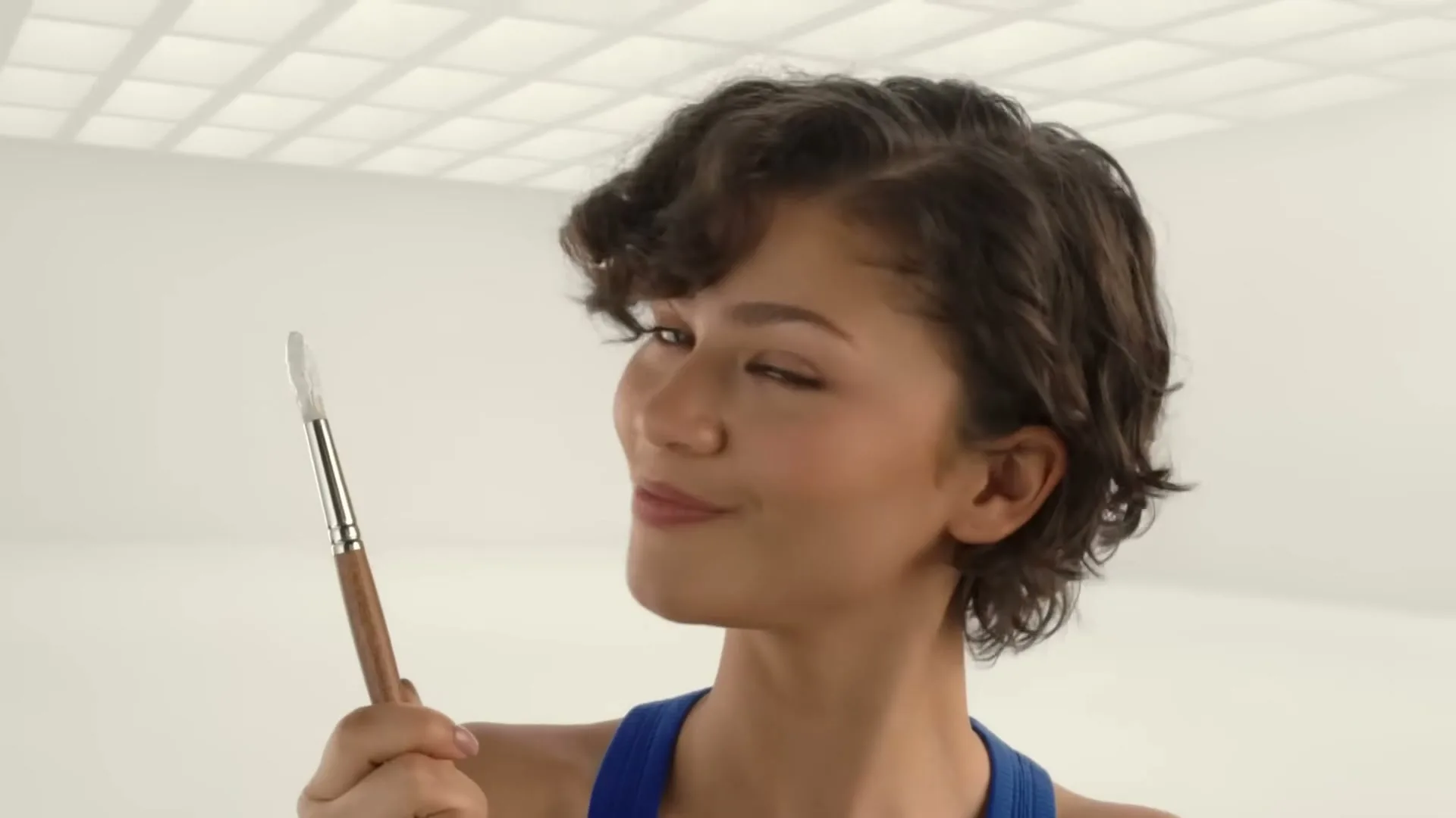 A young woman with short curly hair, wearing a blue top, holds a paintbrush and then punches towards the camera in a white room.