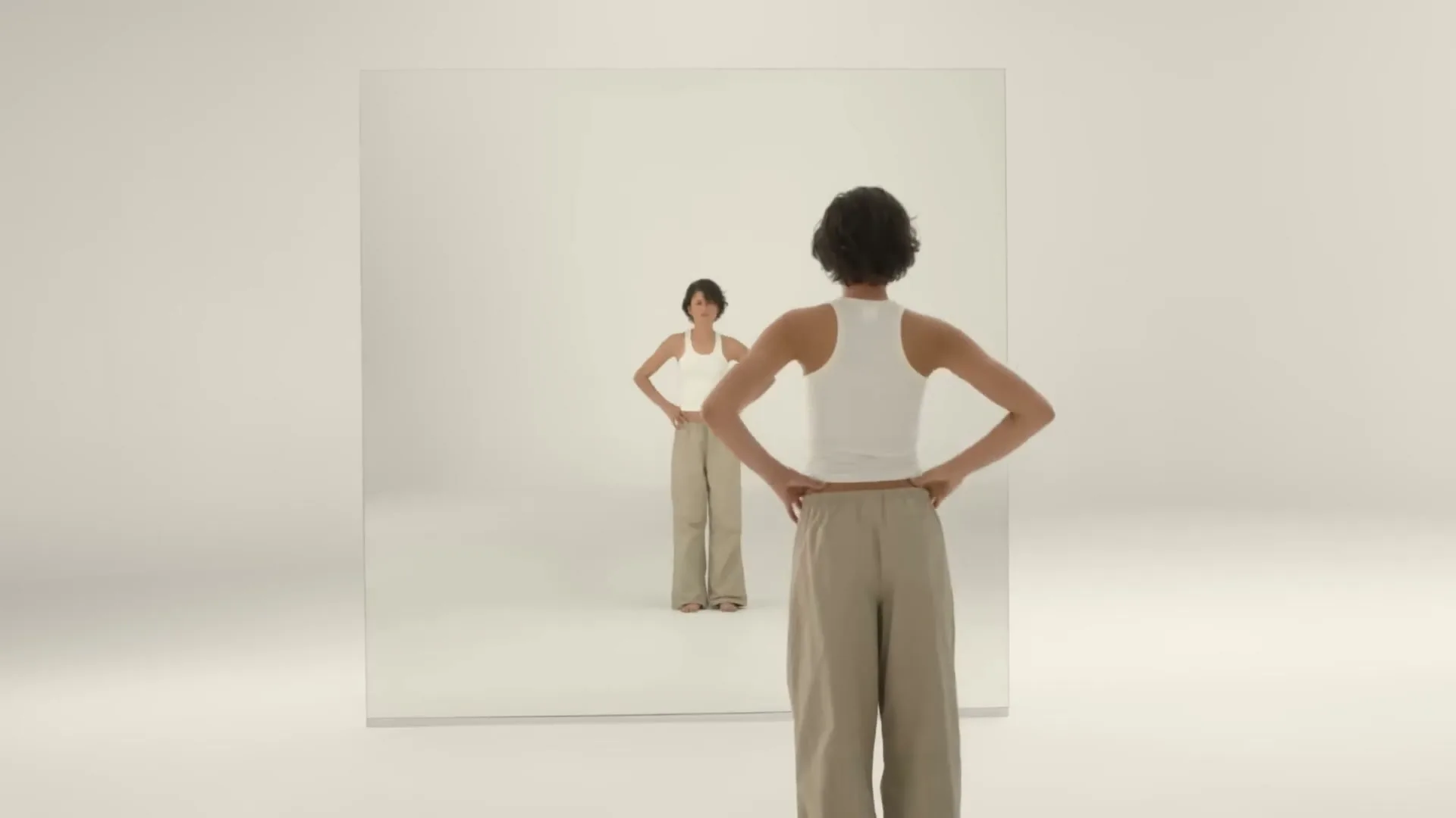 A woman in a blue tank top and white skirt holds a wand, then looks at her reflection in a mirror, then walks away from the camera in a white room.