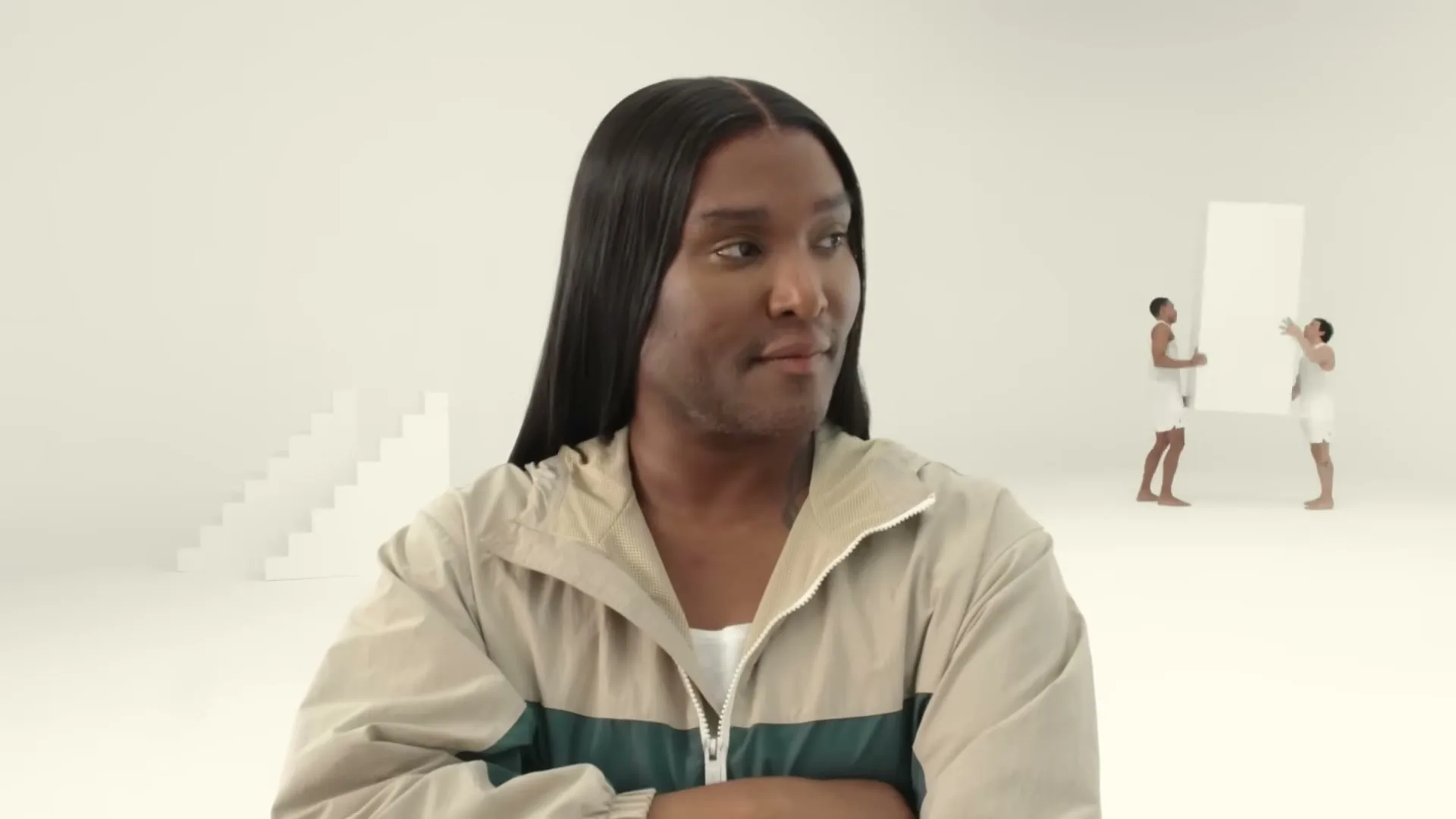 A person with long dark hair and a two-tone jacket looks around a minimalist white space as small figures move white blocks in the background.