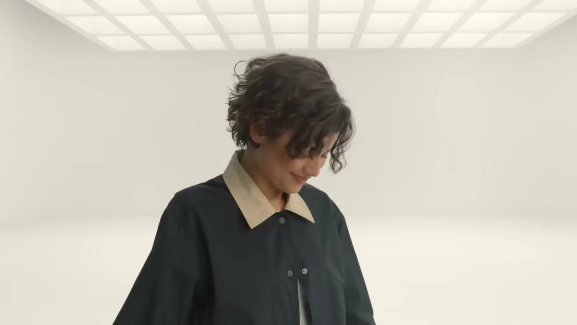 A young woman with short, curly hair, wearing a dark jacket with a light collar, looks around a minimalist white room with grid lighting on the ceiling.