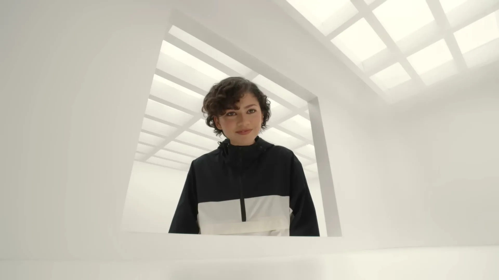 A young woman with short curly hair, wearing a black and white jacket, looks down through a square opening in a white room, then reaches out.