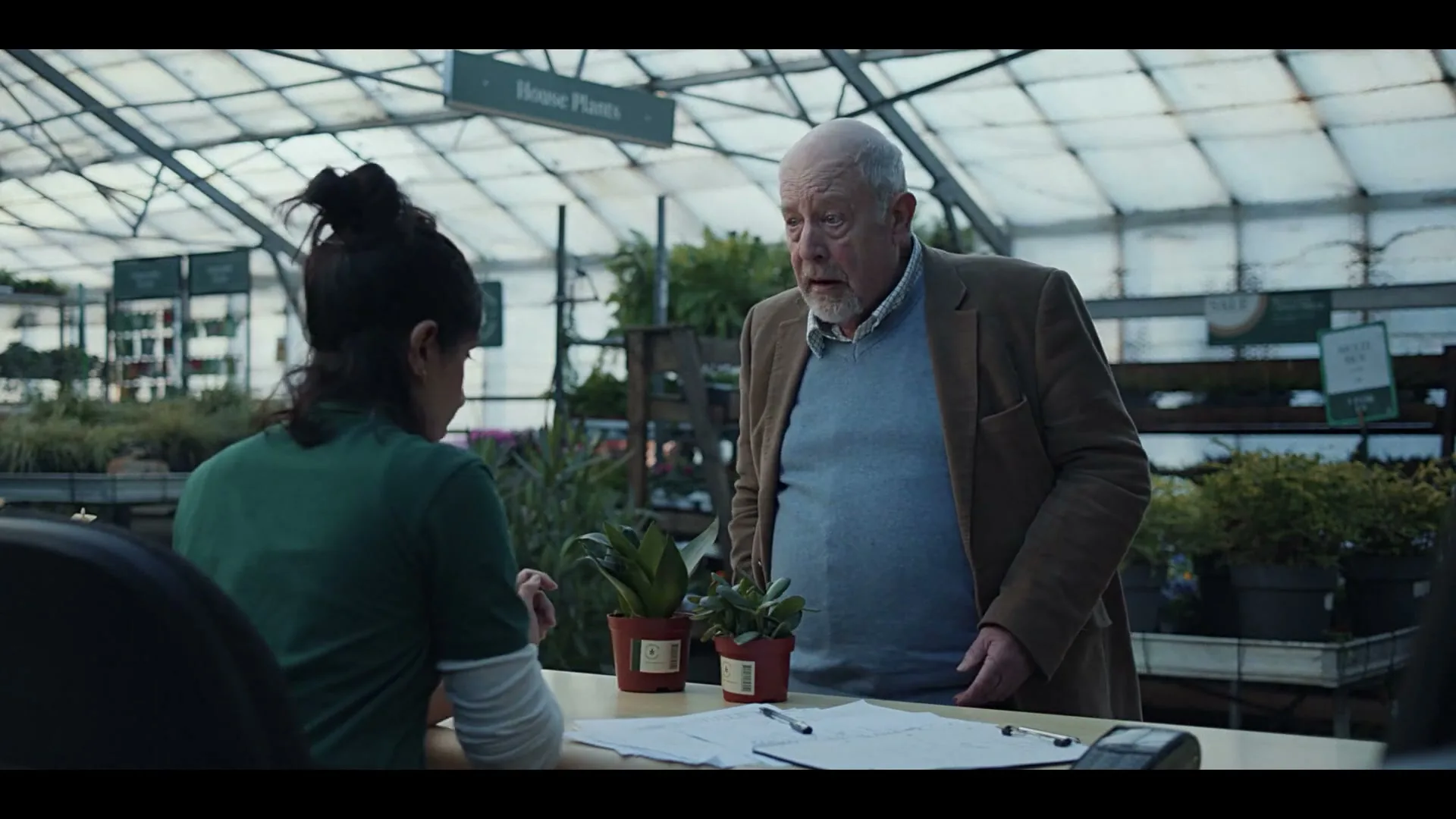 An older man in a brown jacket and blue sweater pays for two small potted plants at a garden center checkout counter, interacting with a female employee.