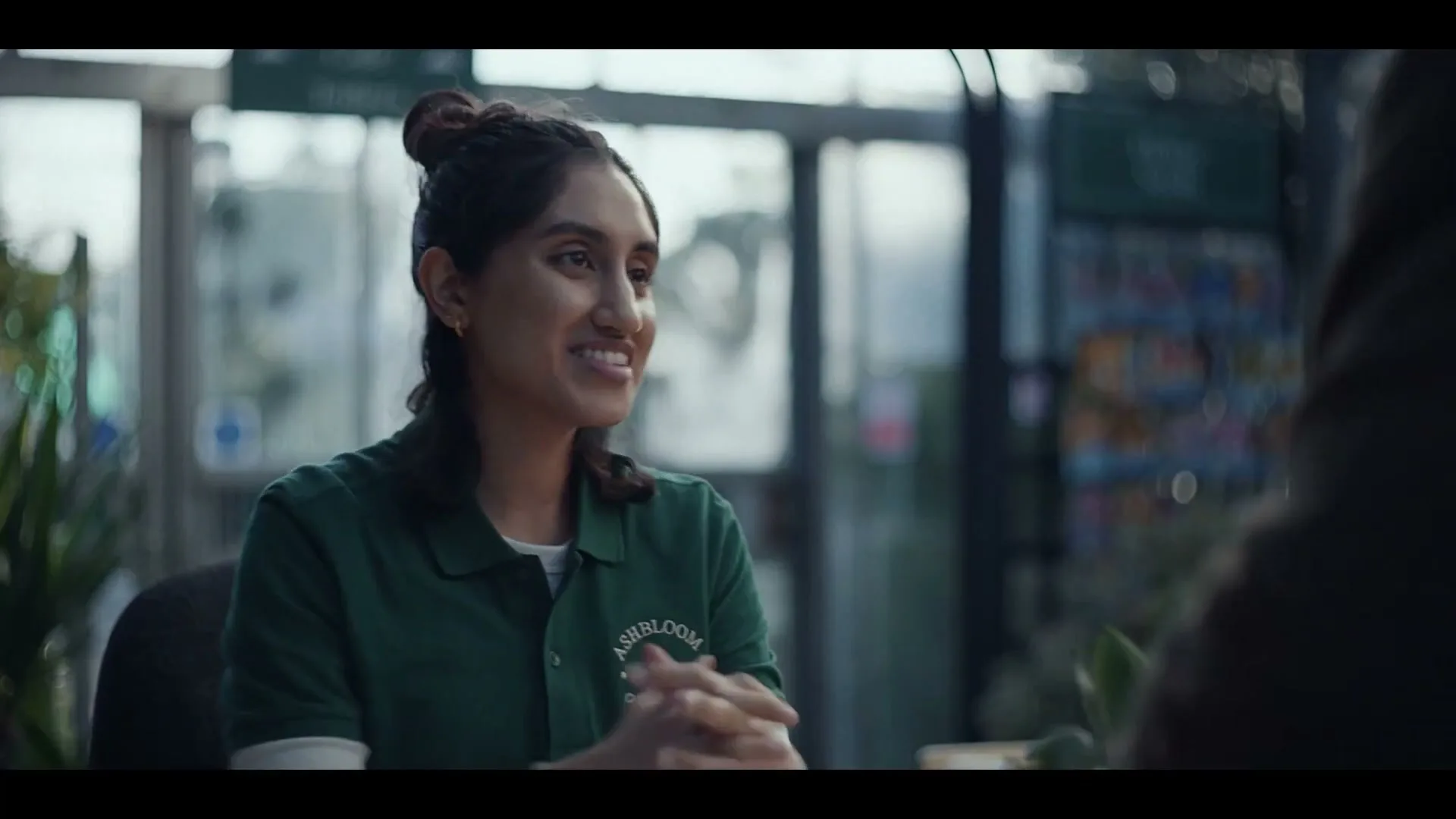 A young South Asian woman with her hair in a bun, wearing a green polo shirt, smiles while holding a card reader at a garden center checkout.