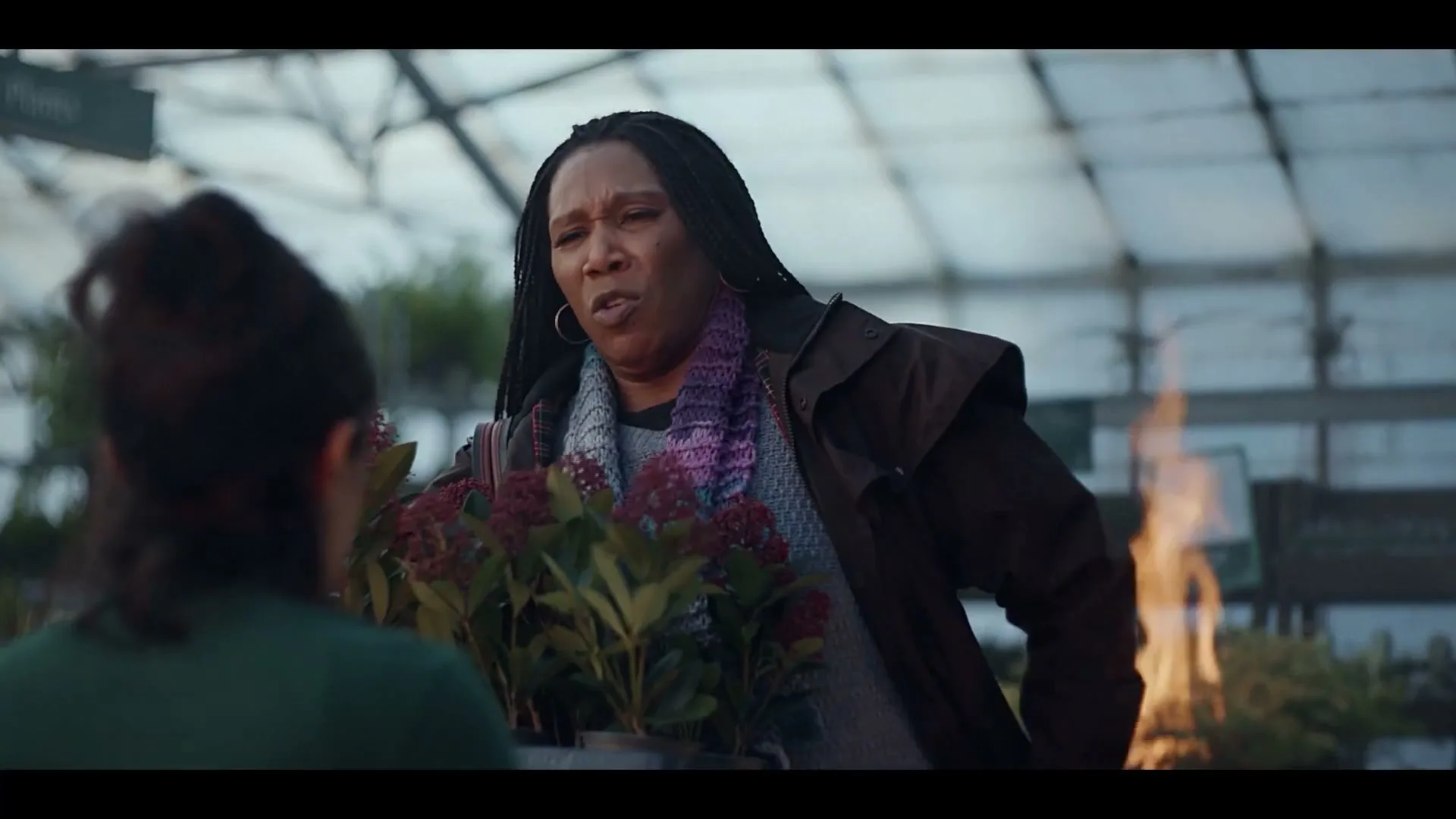 A woman with braided hair and a colorful scarf looks at plants in a greenhouse, talking to another person whose back is to the camera.