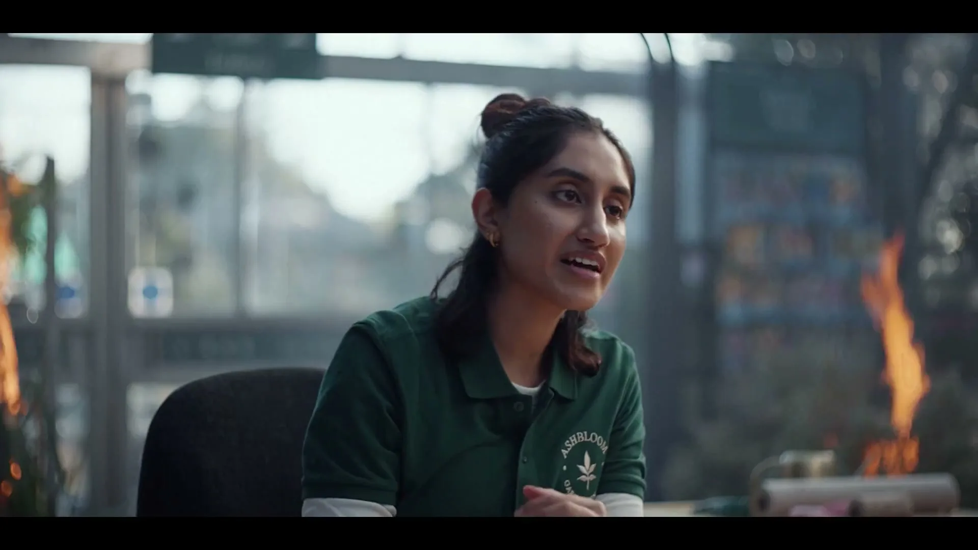 A young South Asian woman with dark hair in a bun, wearing a green polo shirt, sits at a desk in a greenhouse, talking with a concerned expression as fires burn in the background.