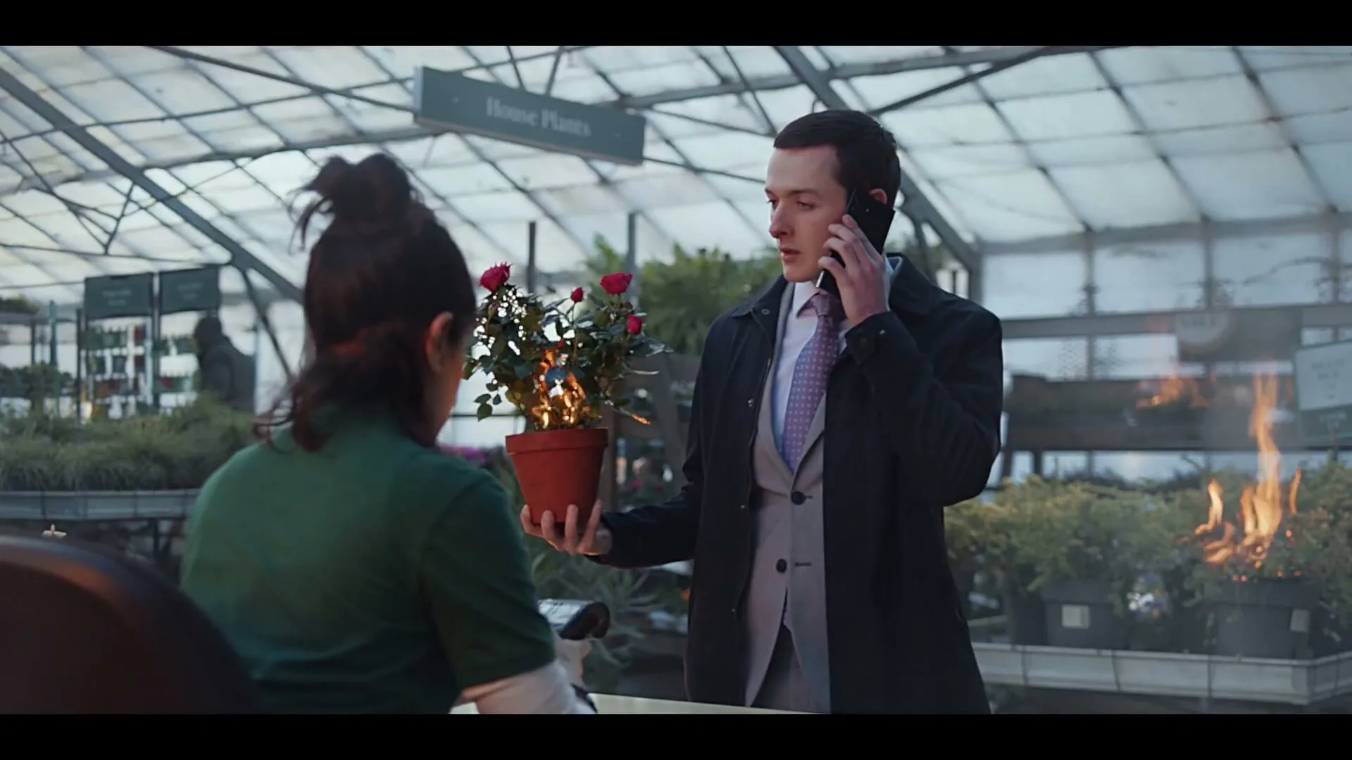 A man in a suit holds a burning potted plant while talking on the phone to a cashier in a greenhouse.