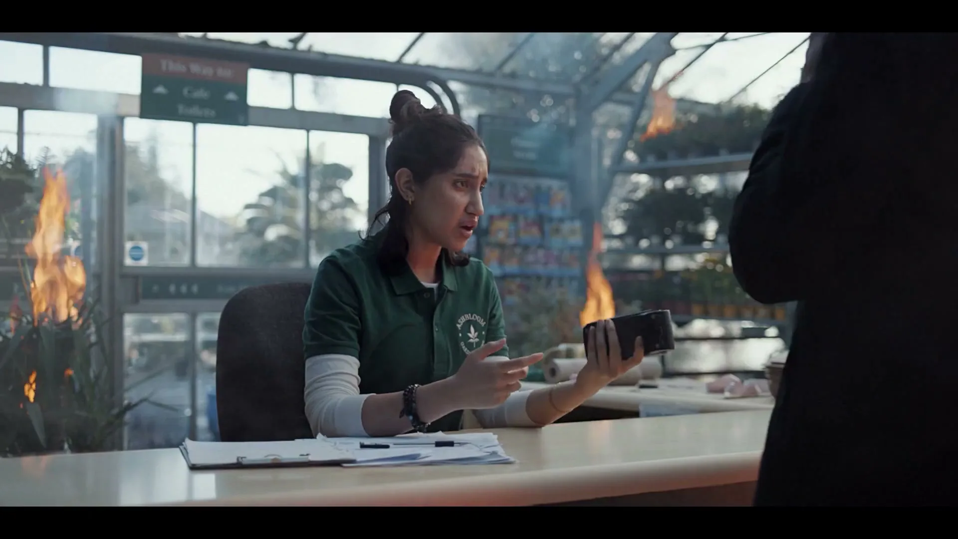 A female cashier in a green uniform holds a card reader, looking confused as plants burn around her in a greenhouse.
